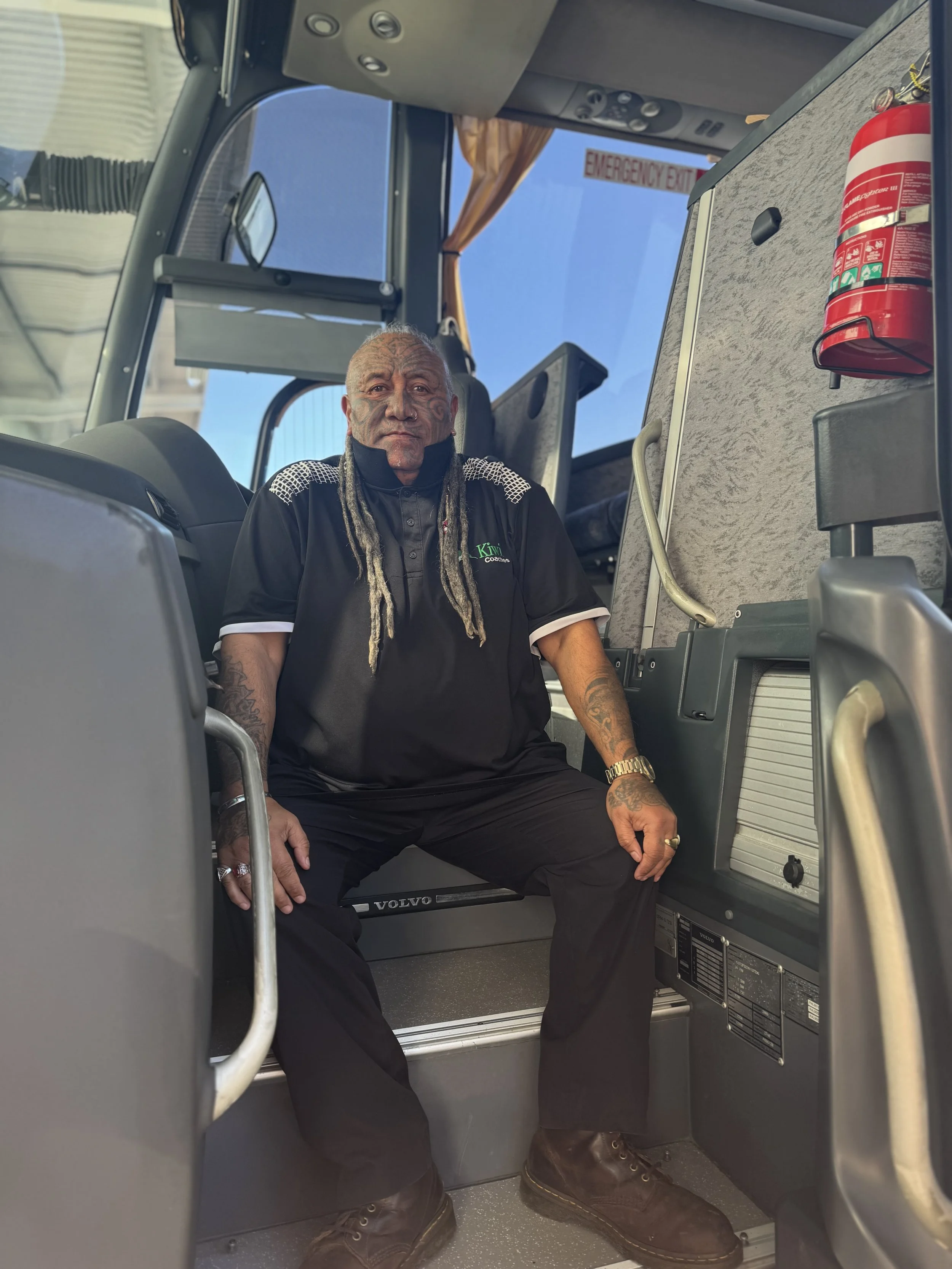 A man with long hair and tattoos sitting inside a bus, with an emergency exit sign visible through the window.