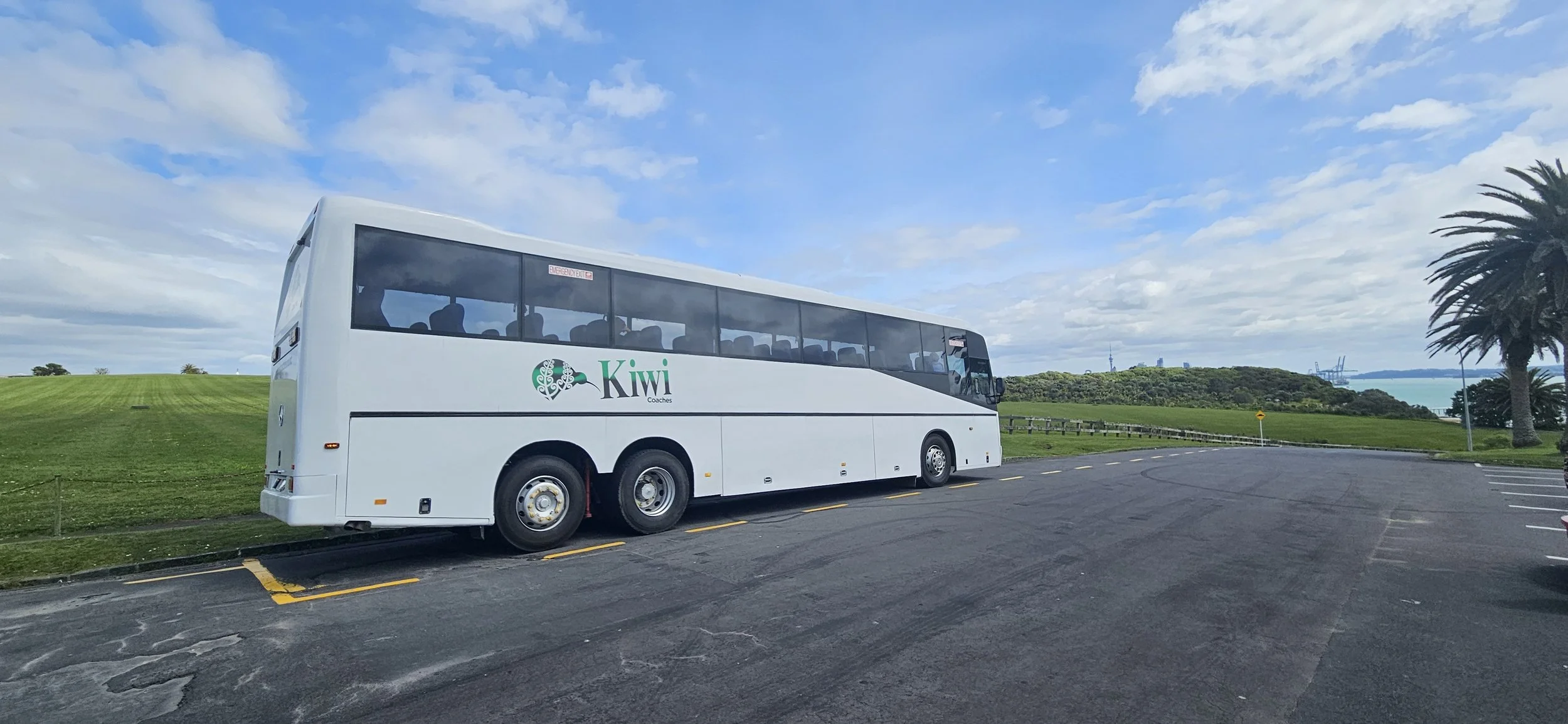 A white coach bus with Kiwi Coaches logo parked in an outdoor parking lot with a grassy landscape, palm trees, and a view of water and hills under a partly cloudy sky.