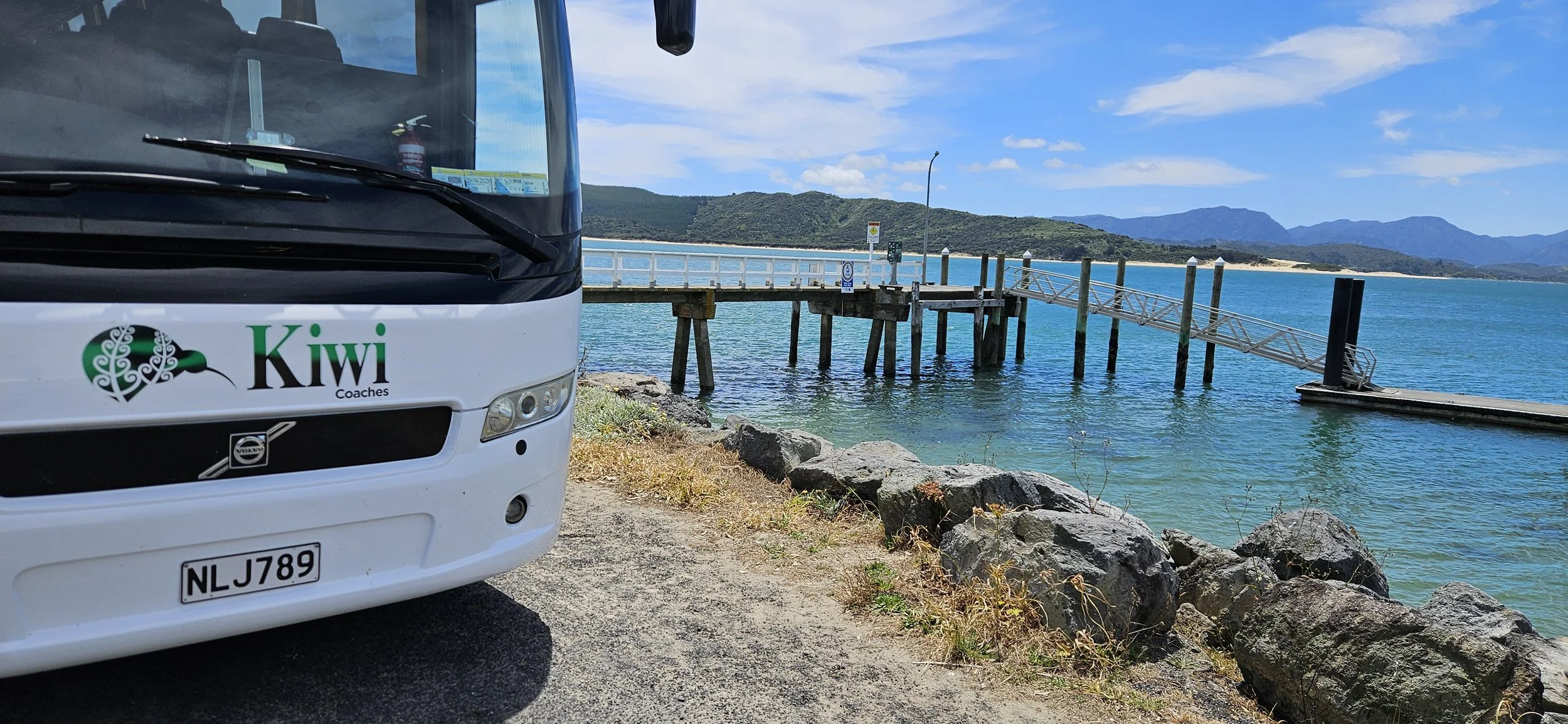 Part of a white bus with a Kiwi Coaches logo on the front, beside a seaside pier with a ramp, rocks, and a calm blue sea with distant hills under a partly cloudy sky.