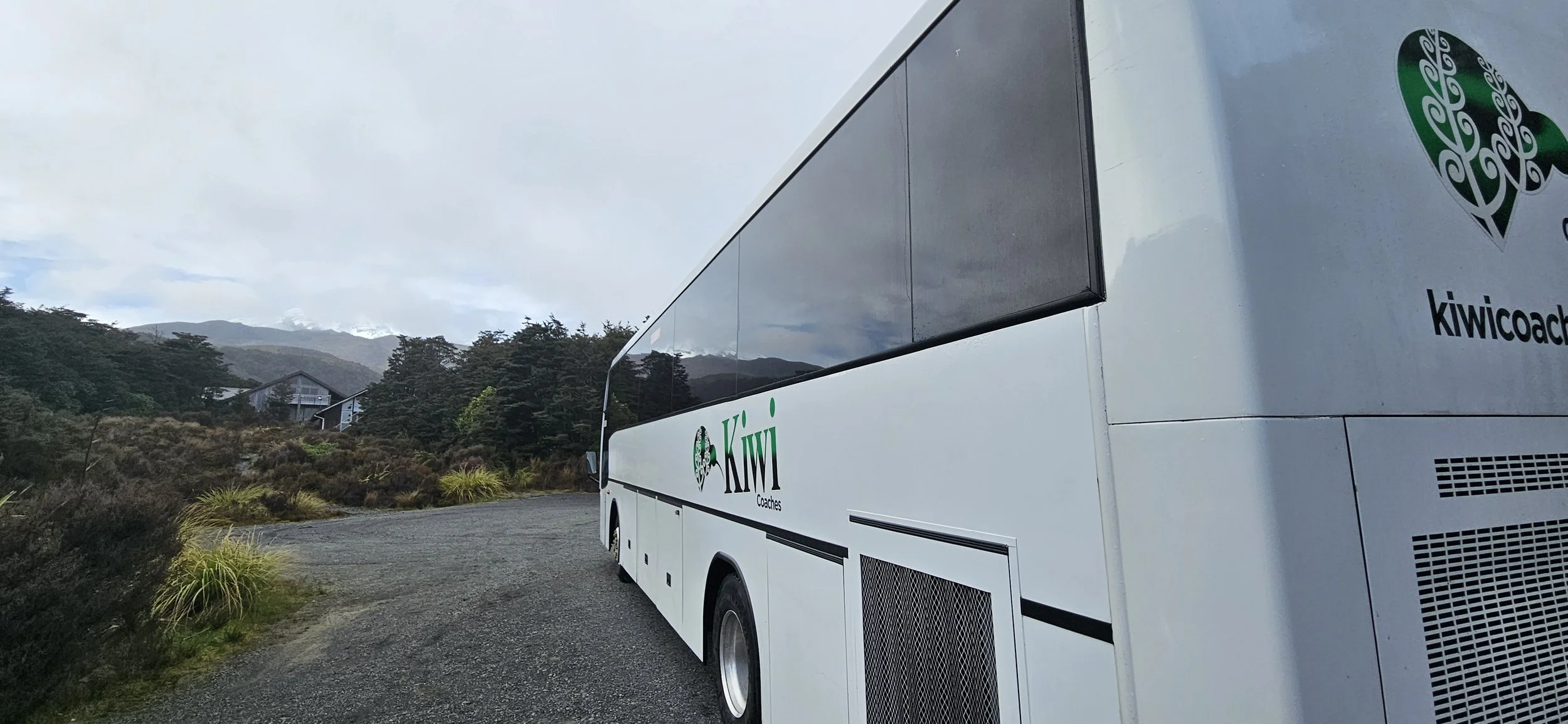 A white bus with Kiwi Coaches logo on the side, parked on a gravel road in a hilly, forested area with mountains in the background.