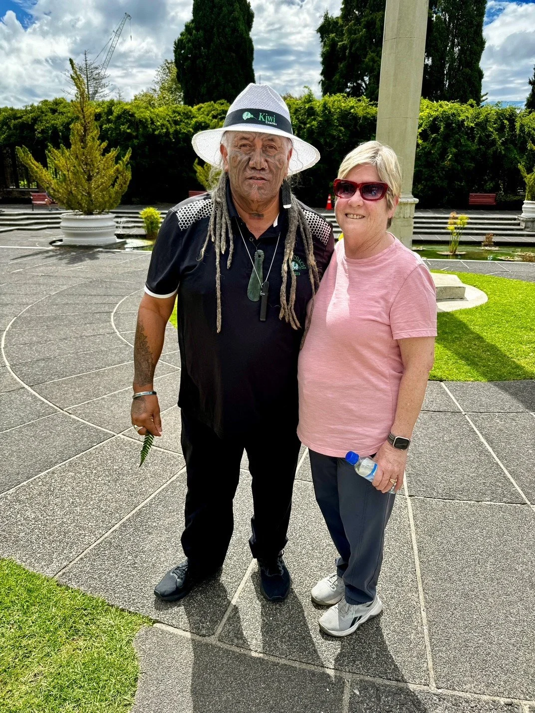 A man with dreadlocks and a woman with short hair and sunglasses standing together outdoors on a paved area, with greenery and trees in the background.