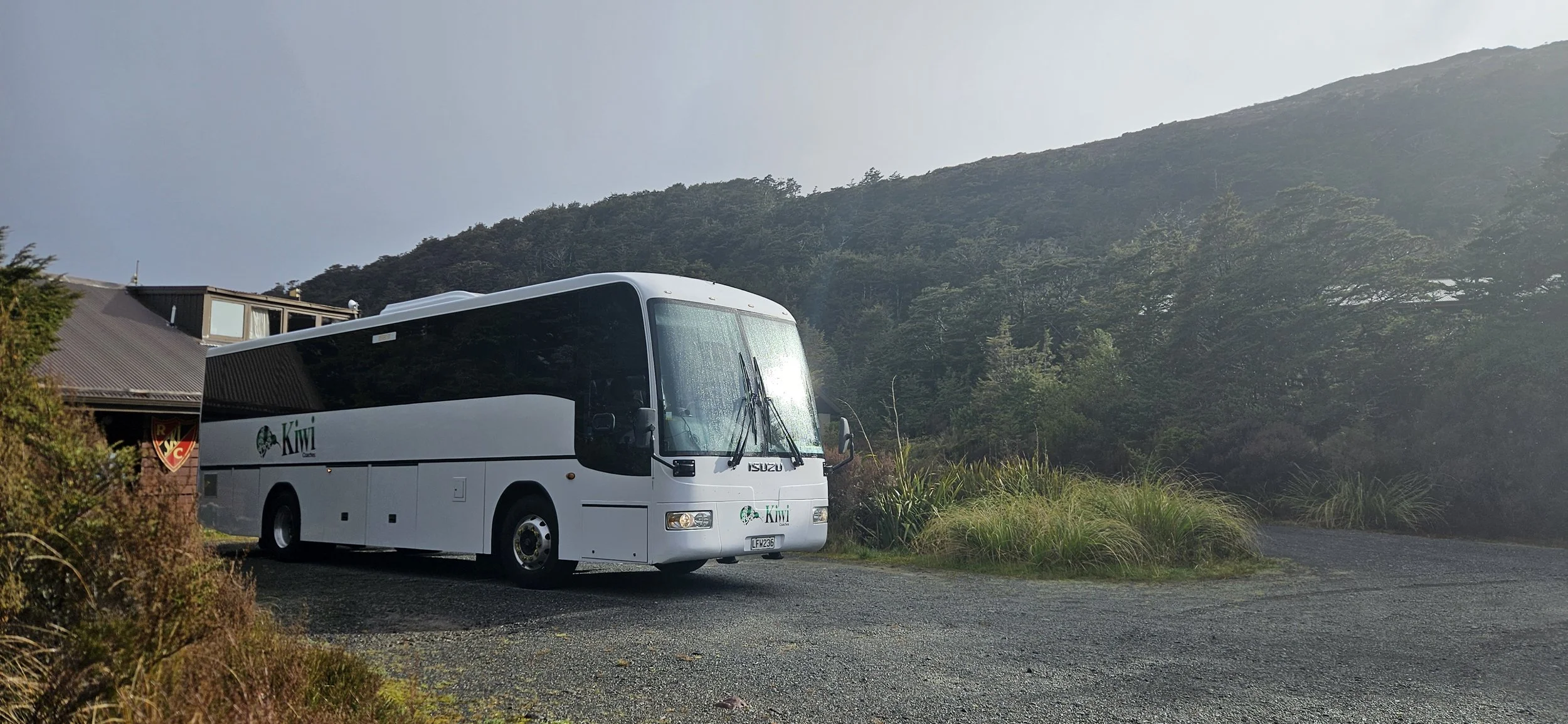 A white bus with Kiwi Tours branding parked on a gravel path near lush green vegetation and a forested hillside under a partly cloudy sky.