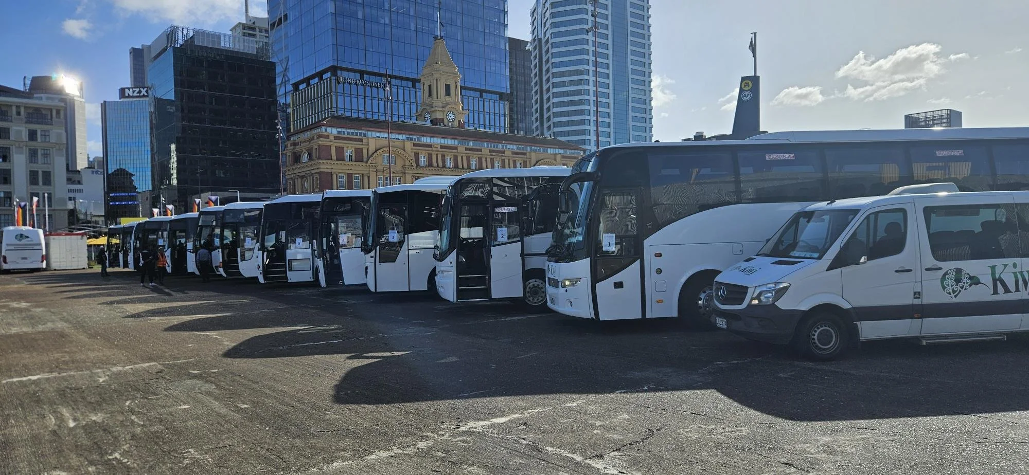 Several white buses parked in a row in an urban area with tall buildings and a historic clock tower in the background, under a cloudy sky.