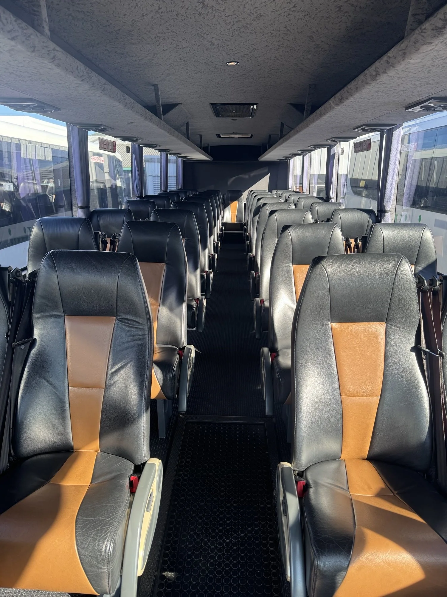 Empty bus interior with black and brown leather seats, large windows on both sides, and sunlight streaming through.