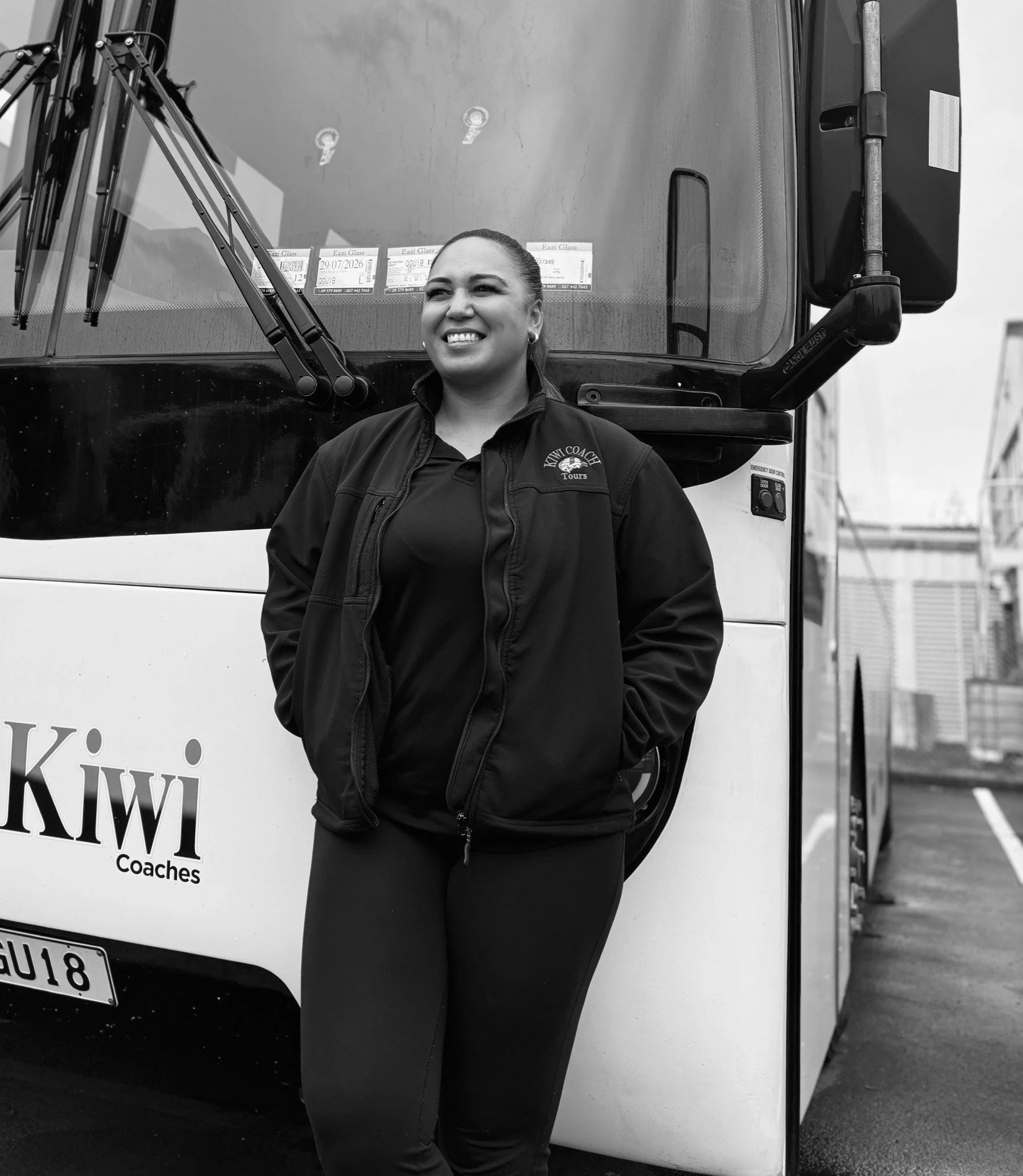 A smiling woman in a uniform standing in front of a bus with "Kiwi Coaches" branding, outdoors on a paved lot.