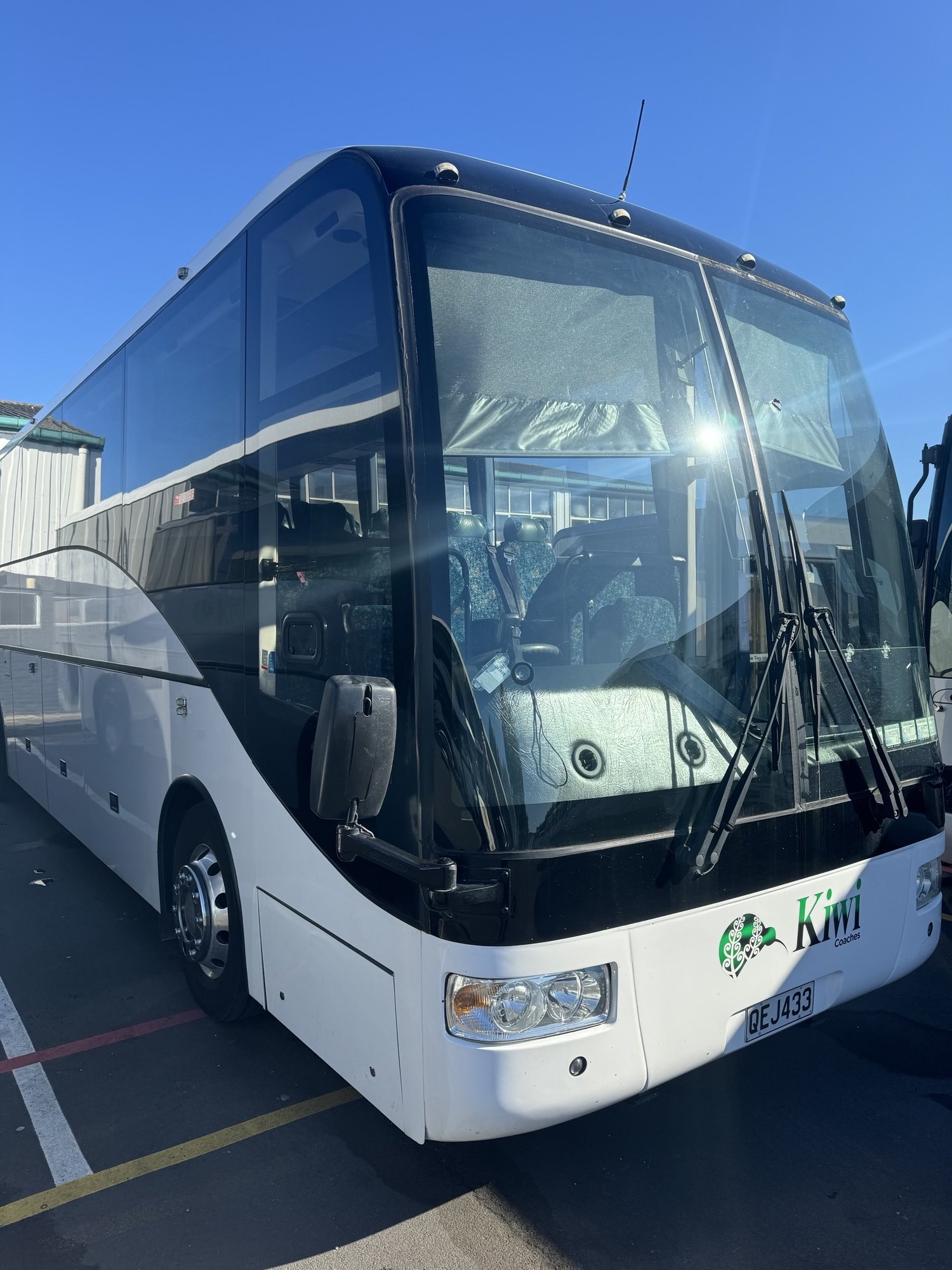 A white and black tour bus with the logo 'Kiwi Coaches' parked on a street under a blue sky.