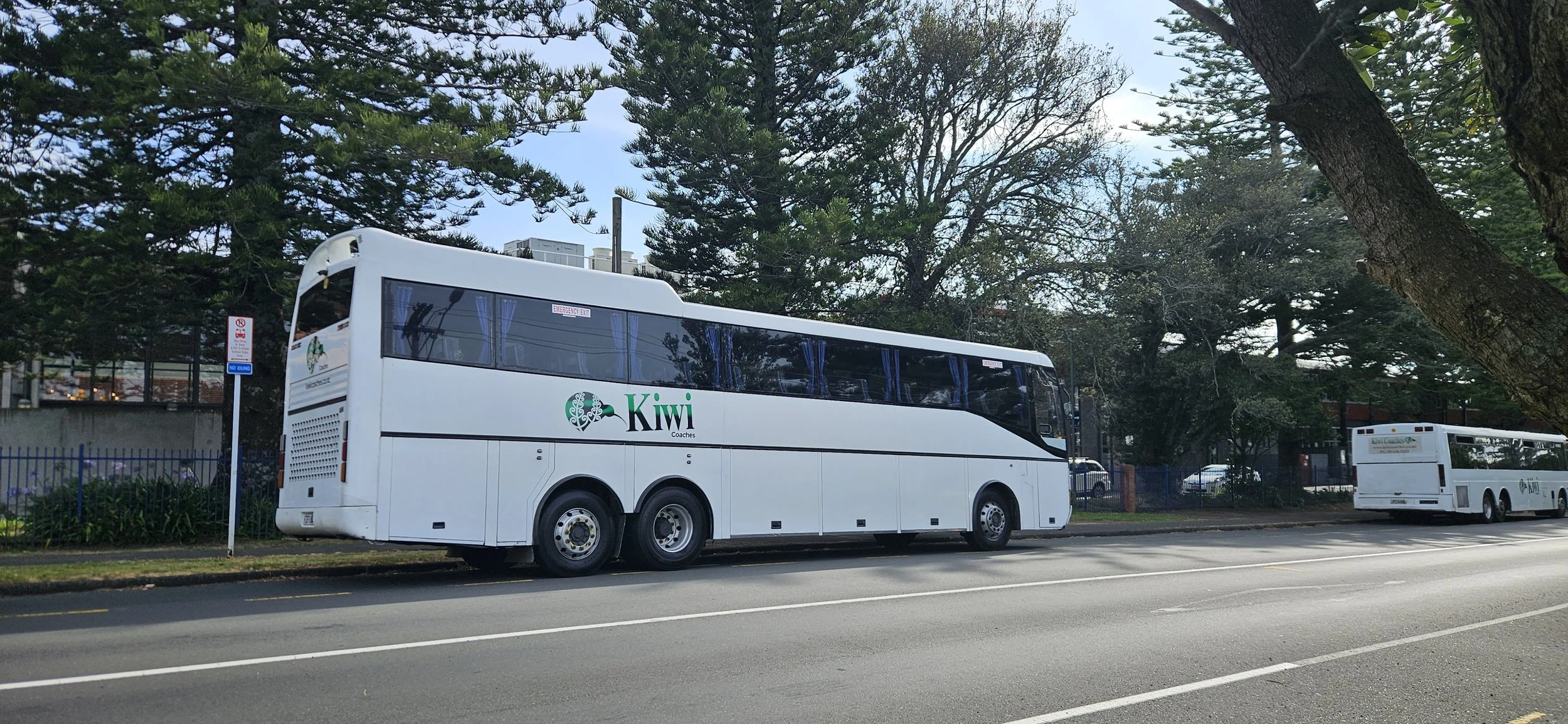 White double-decker coach bus with Kiwi Coach logo parked by the side of the road, trees in the background, and a no parking sign nearby.