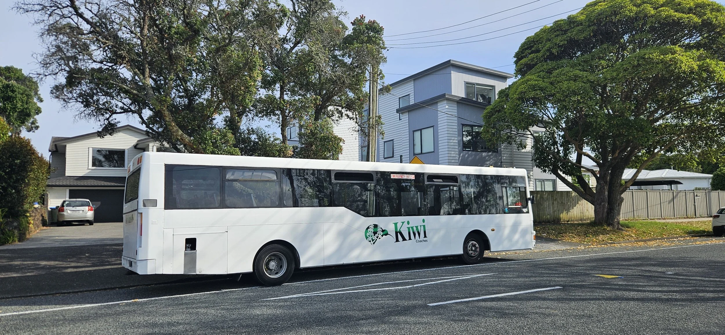 White Kiwi Coaches bus parked on the side of a street near residential houses and trees.