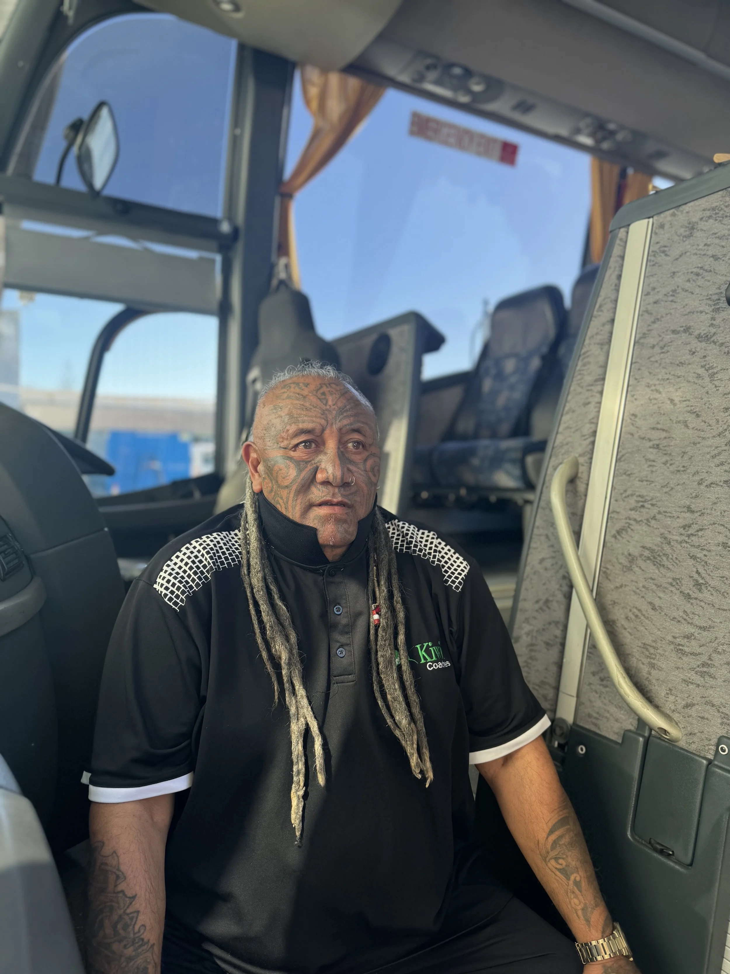 A man with tattoos and long gray dreadlocks sitting inside a bus with yellow curtains and seats, looking thoughtful.