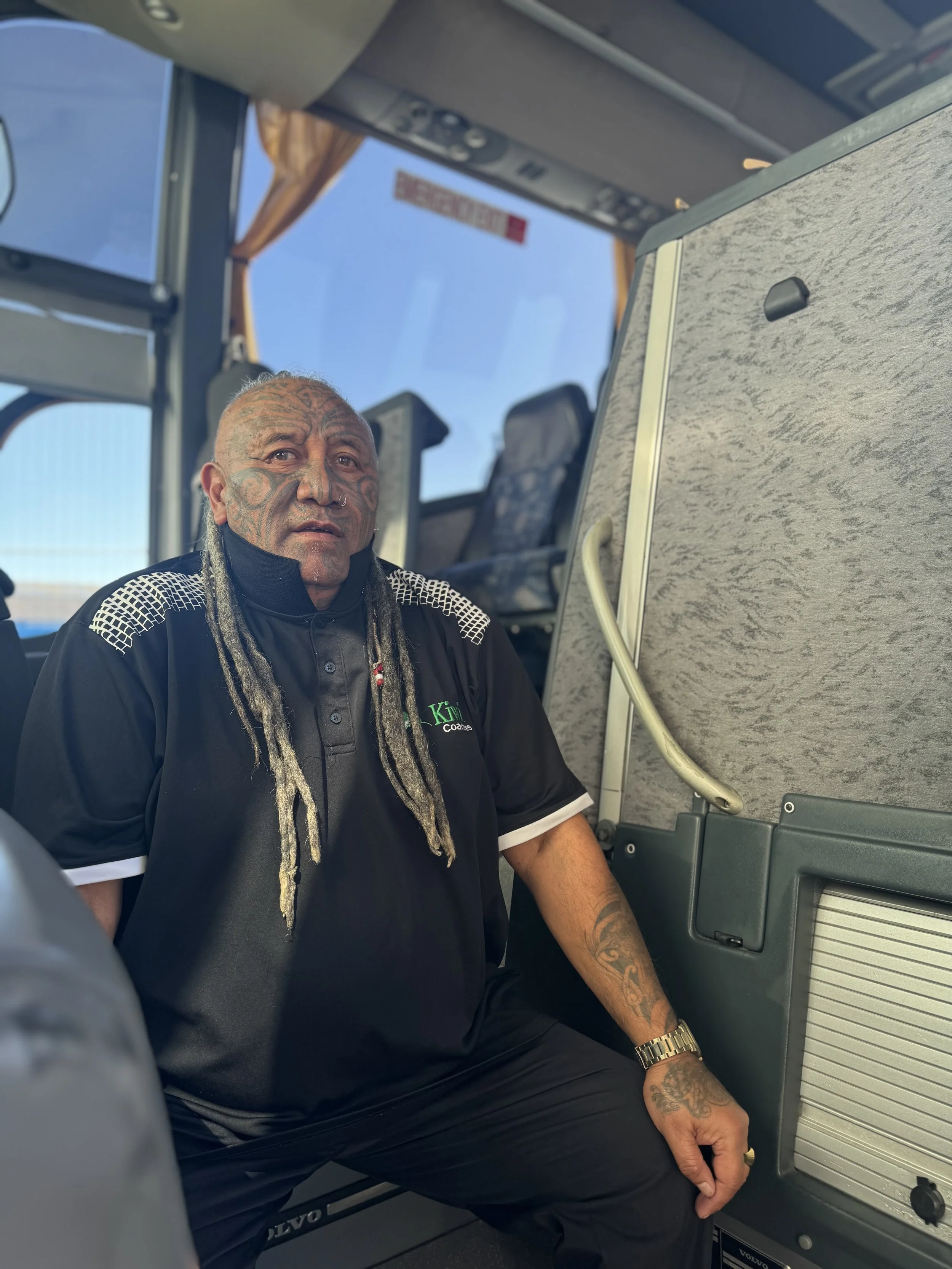 A man with tattoos and dreadlocks sitting inside a bus or coach, looking at the camera.
