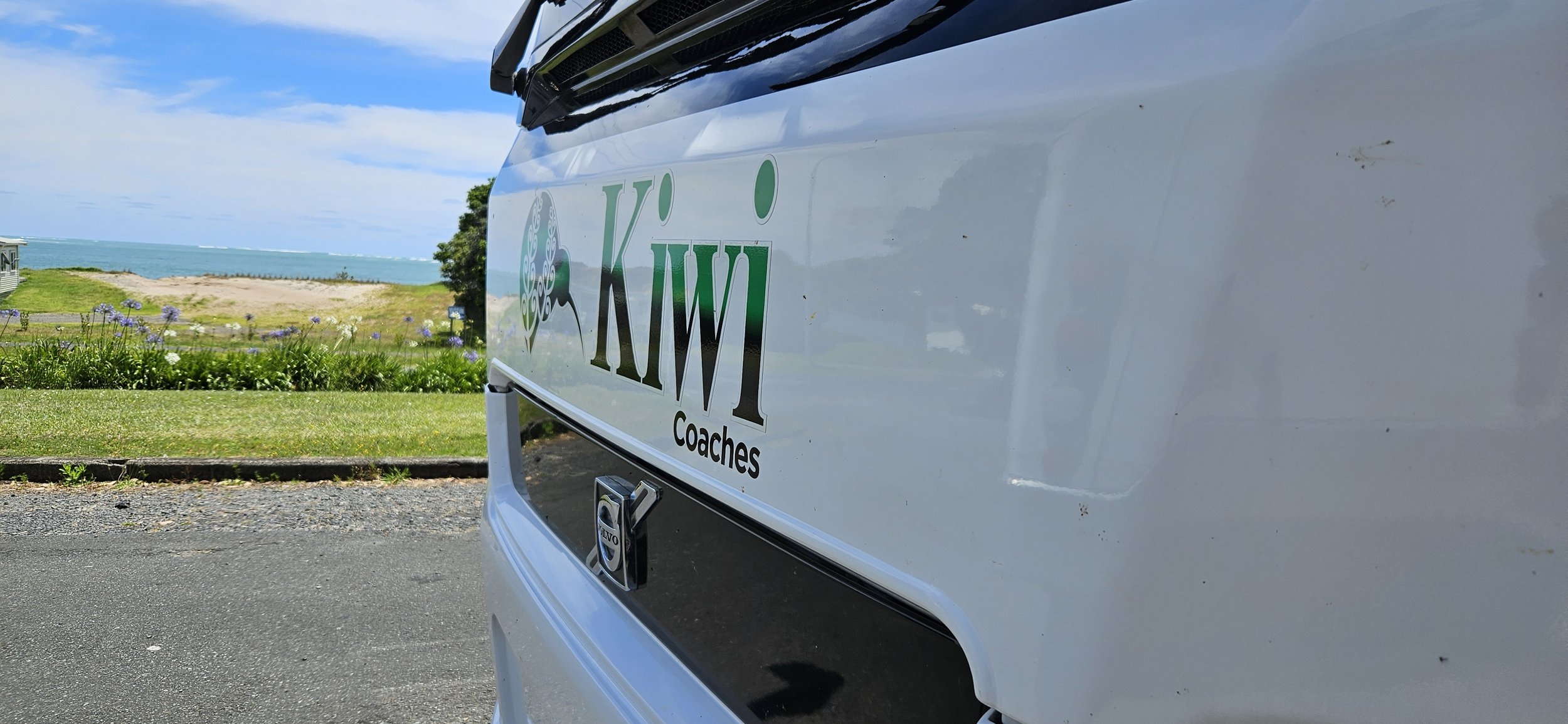 Close-up of a white vehicle with a logo that reads "K&W Coaches" and a Volvo emblem, parked near a grassy area with flowers, a beach, and the ocean in the background during daytime.
