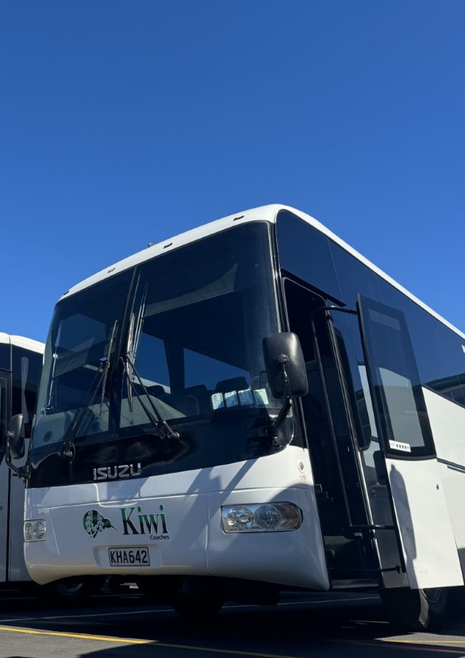 White bus with Kiwi Coaches logo parked in a lot under a clear blue sky.