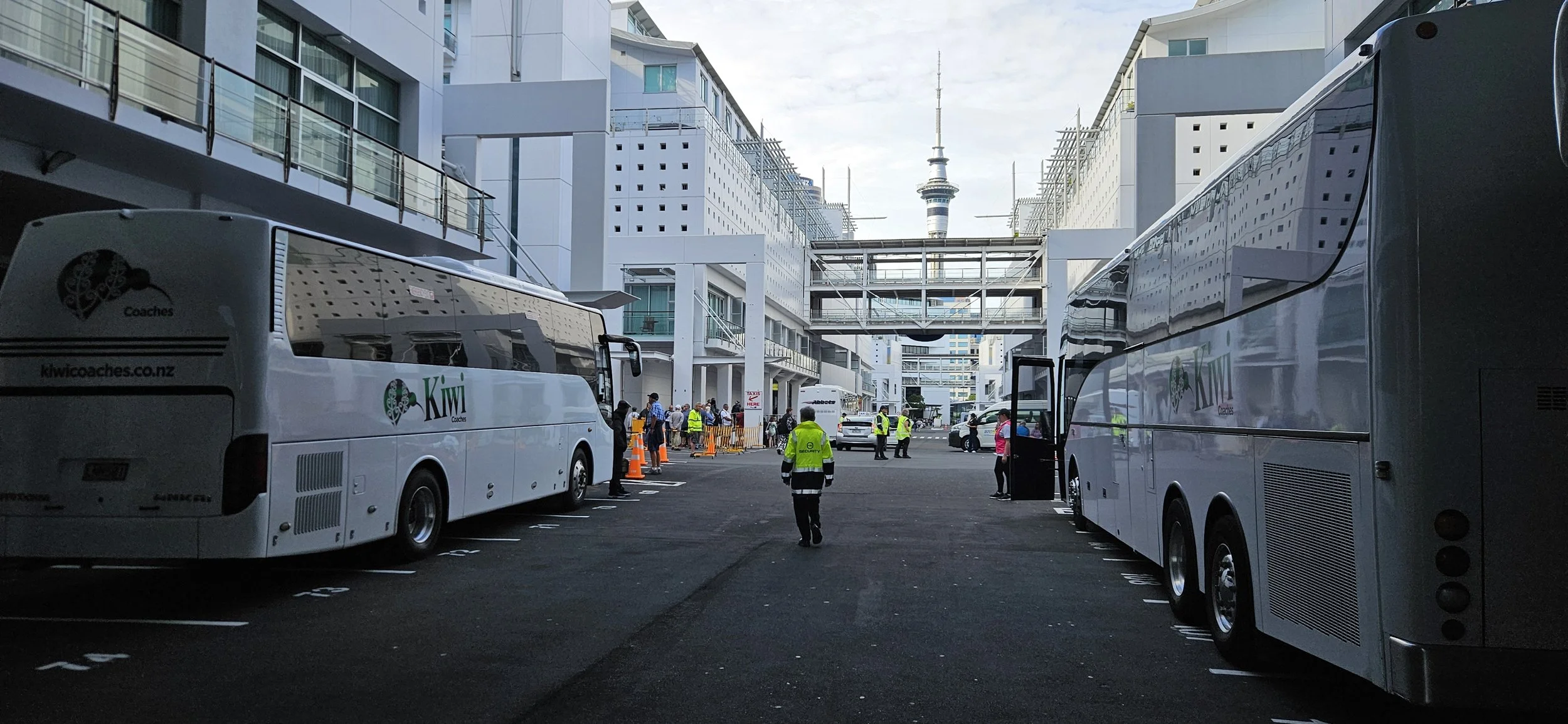 Parking lot with two large Kiwi Coaches buses on either side, several people in safety vests and casual clothes, and a few cars in the background. Modern buildings and a tower are visible in the distance, under a cloudy sky.