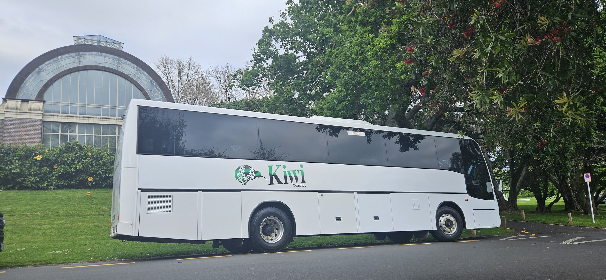 White bus with 'Kiwi Coaches' logo parked on the side of a road amidst green grass and trees, with a building featuring arched glass windows in the background.