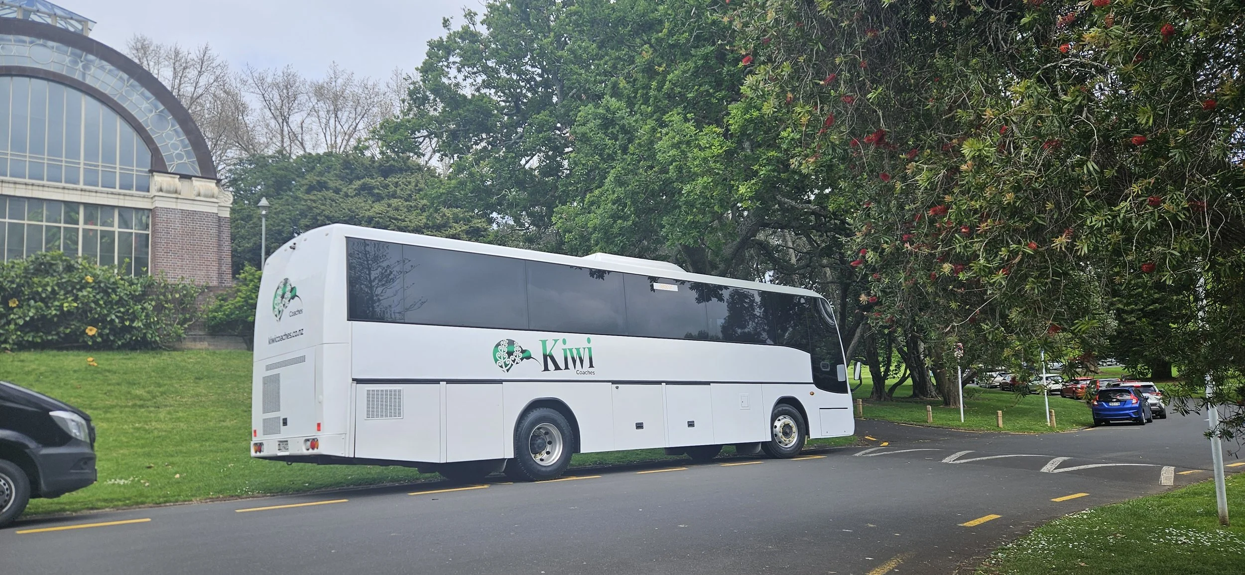 White coach bus labeled 'Kivi Coaches' parked along a road surrounded by green trees and grass, near a brick and glass building.