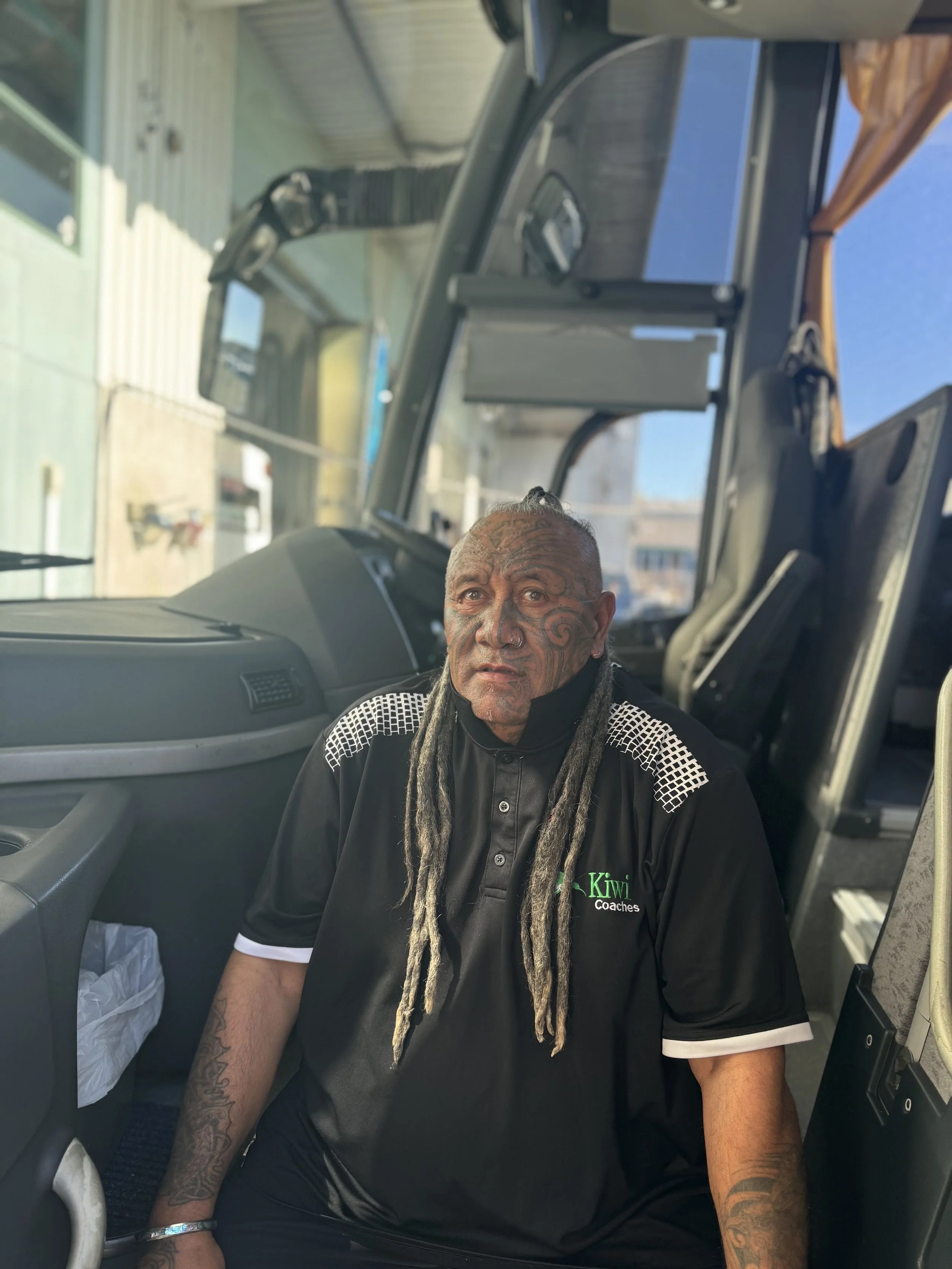 A man with tattoos and dreadlocks sitting inside the driver's seat of a bus, wearing a black collared shirt with the logo 'Kiwi Coach'.