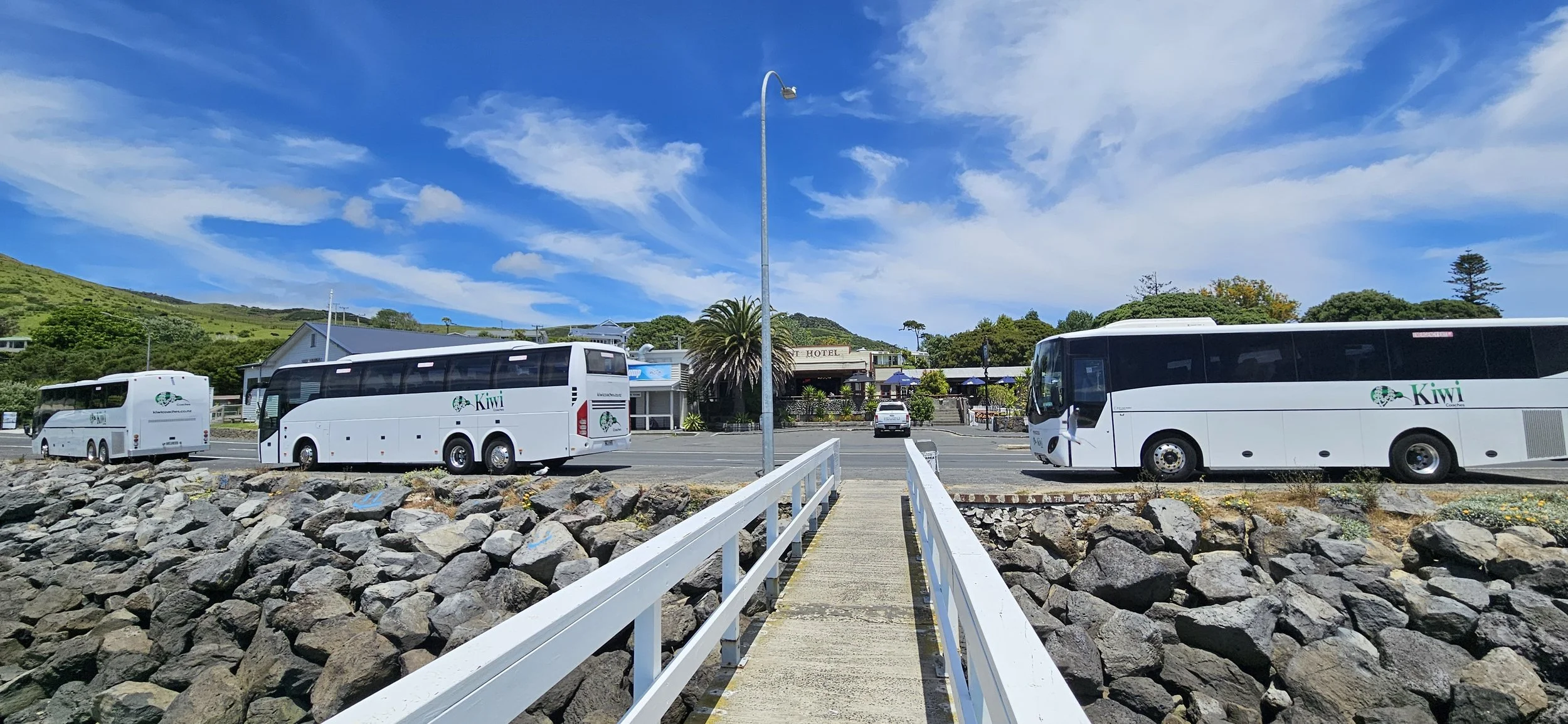 Pedestrian bridge leading to three white coach buses with green Kiwi logo parked on the street. A hotel and green hills are in the background under a blue sky with scattered clouds.