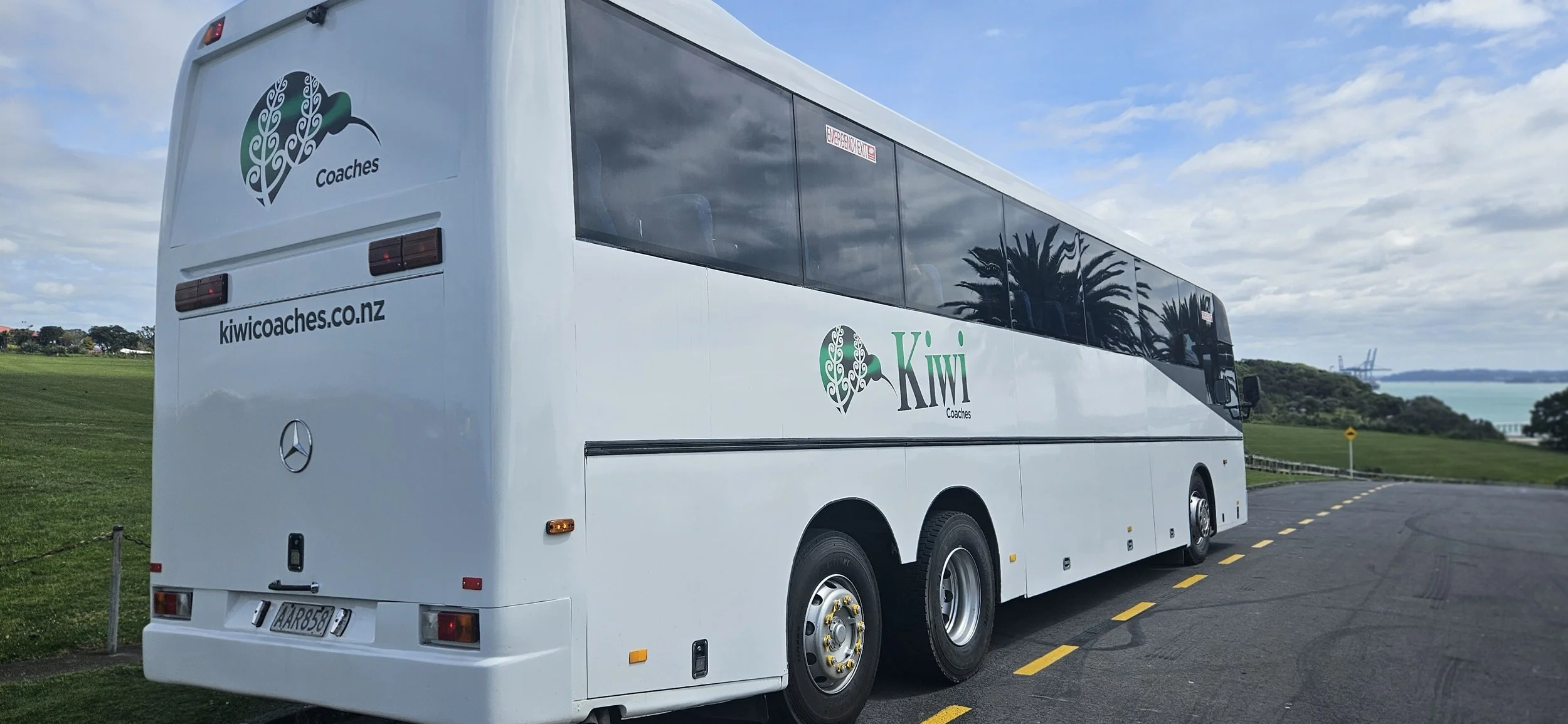 A white coach bus with 'Kiwi Coaches' logo and website, parked on a road near green fields with trees, palm trees, and a body of water in the background under a partly cloudy sky.