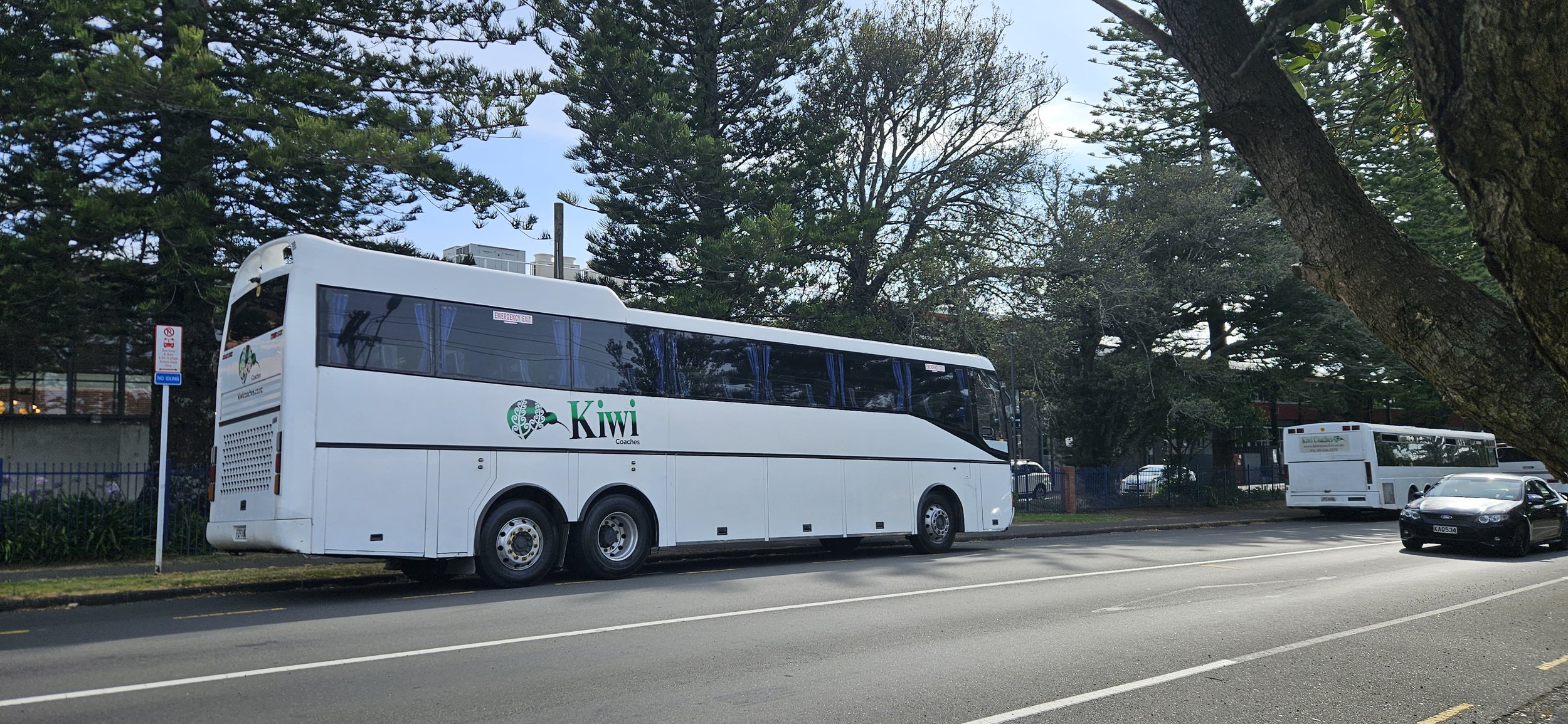 A white Kiwi Coaches bus parked on a street lined with trees, with a black car and another bus nearby, under a partly cloudy sky.