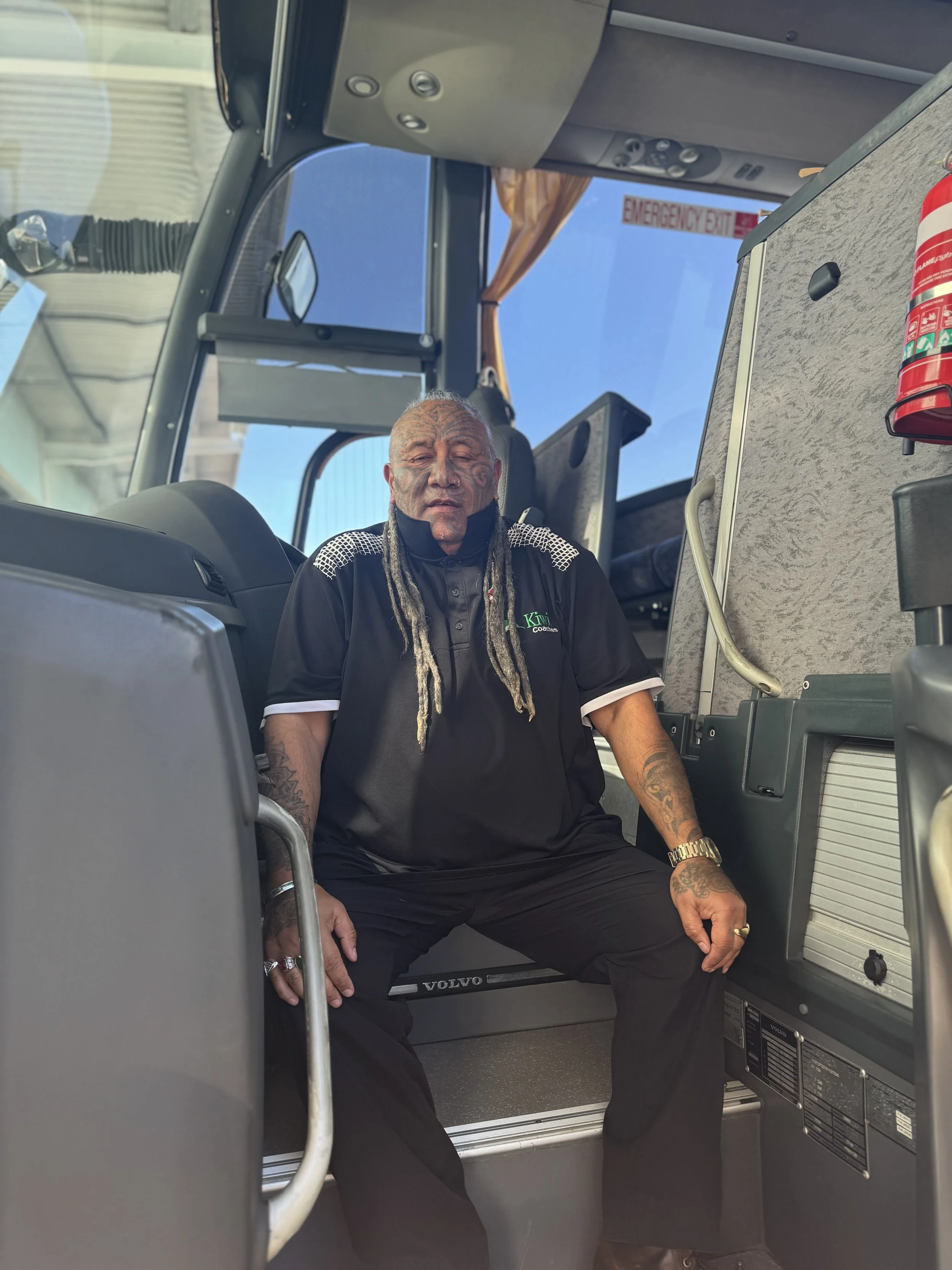 A man with tattoos and dreadlocks sitting inside a bus cockpit.