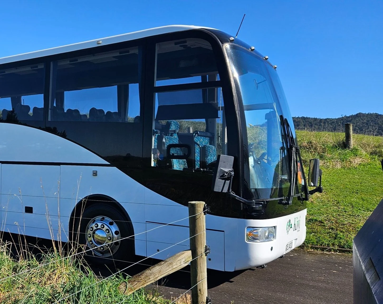A white tour bus parked on a road next to a grassy field, under a clear blue sky.