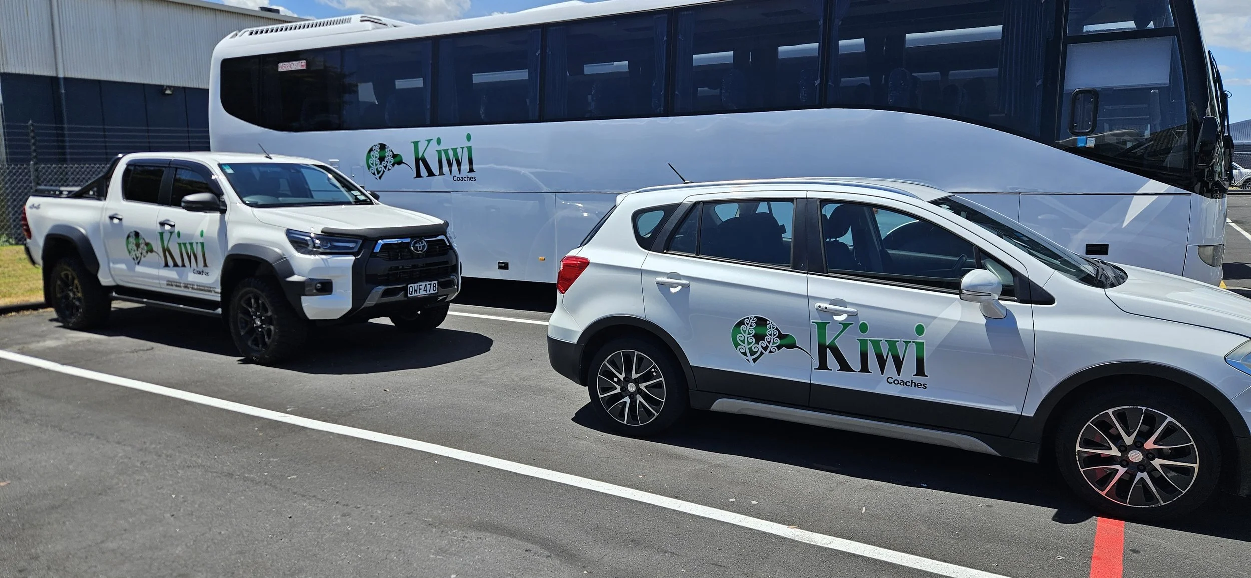 Parking lot with a white Toyota Hilux pickup truck and a white hatchback, both branded with Kiwi Coach logo, parked near a large white bus with the same logo.