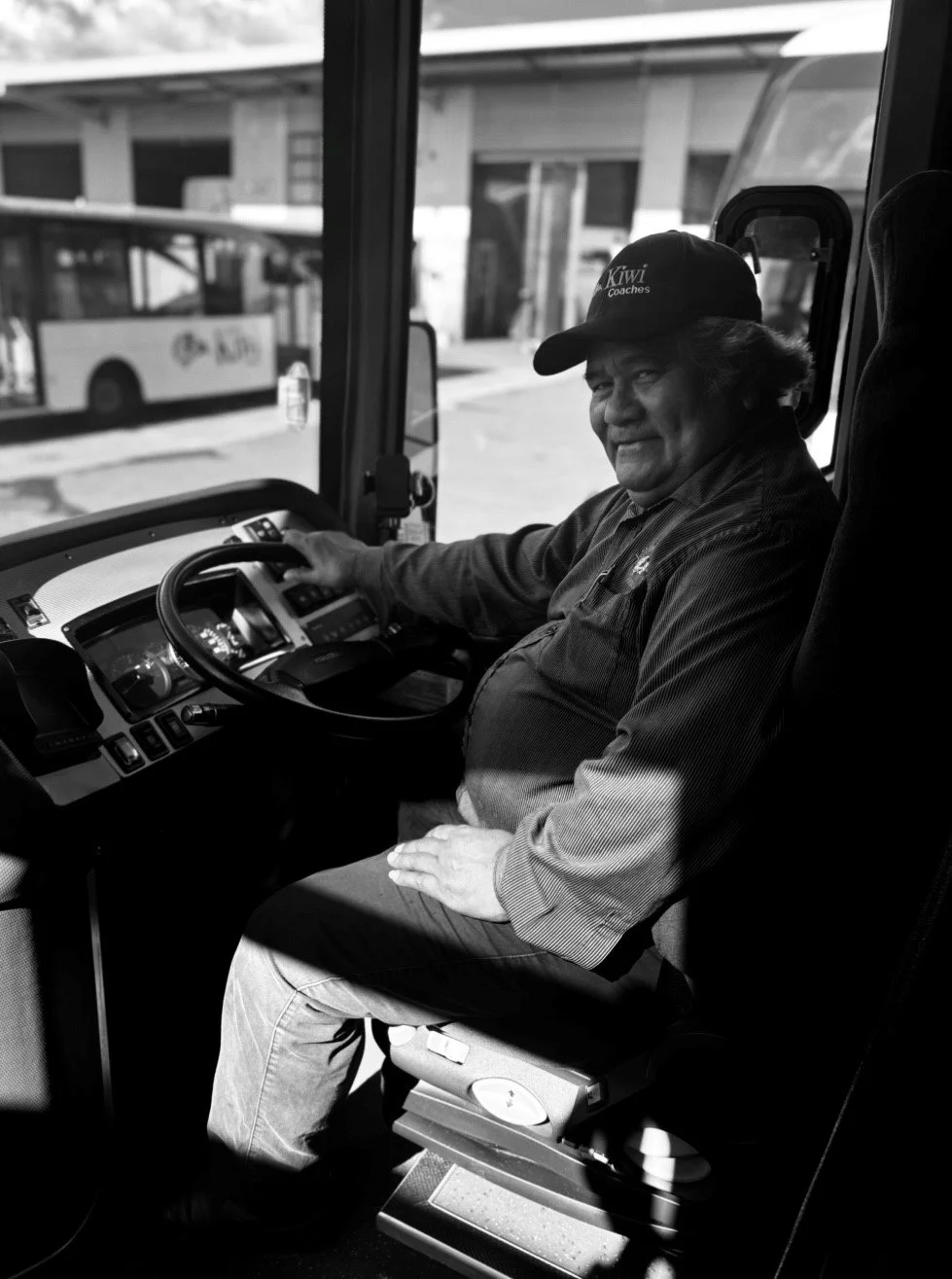 A man sitting in the driver's seat of a bus, wearing a cap that reads 'Kiwi Coaches,' smiling at the camera, with buses and buildings visible through the window behind him.