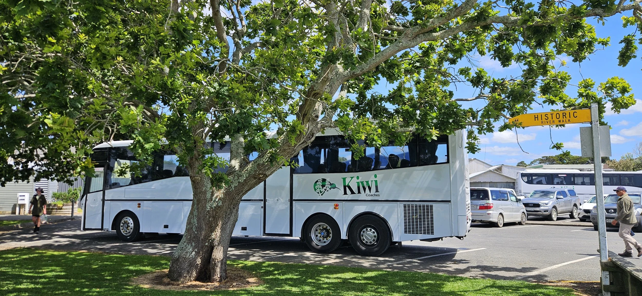 A white Kiwi coach bus parked in a lot near a tree, with several cars behind it. A yellow street sign labeled "HISTORIC RIVER WALK" is visible to the right. People are walking in the parking lot on a sunny day with a blue sky.