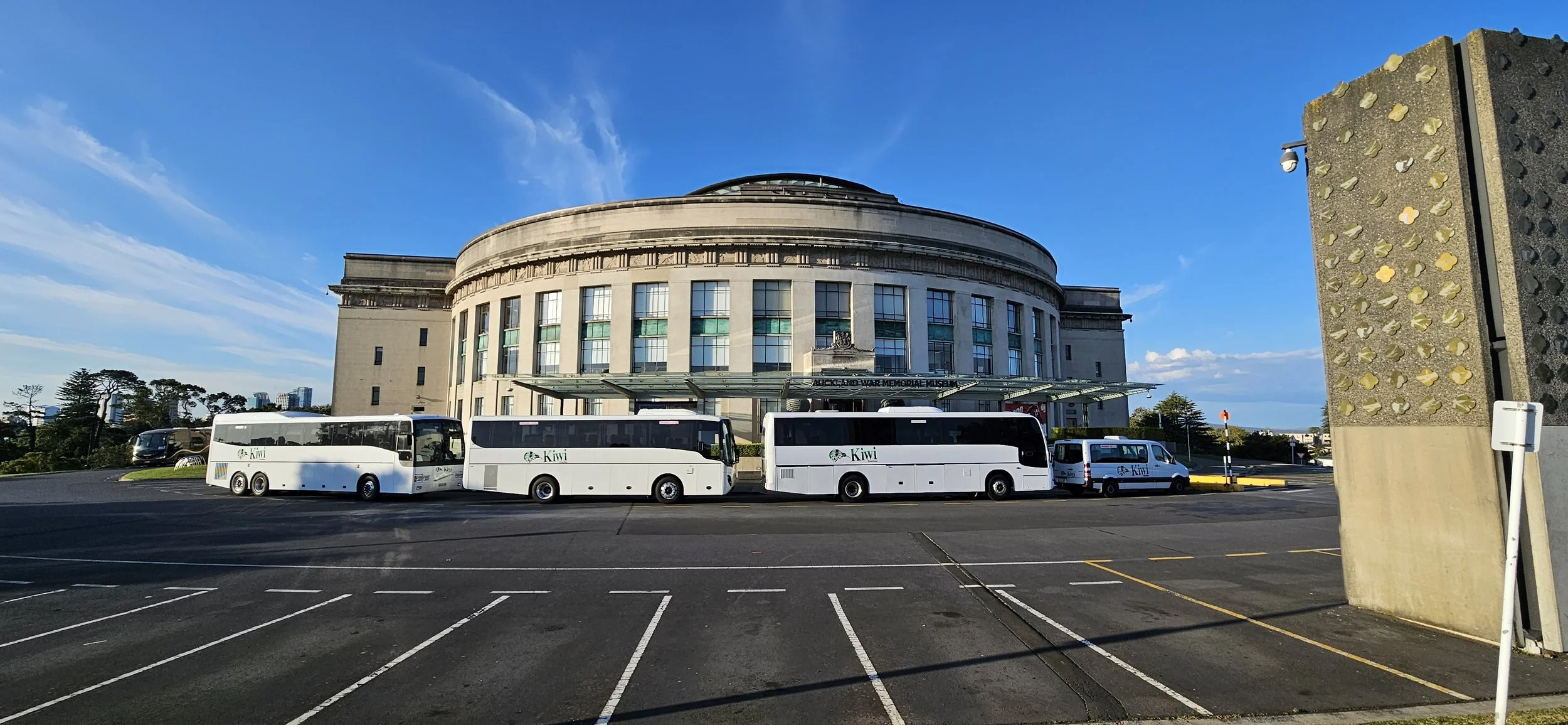 The Auckland War Memorial Museum in Auckland, New Zealand, with four white buses parked in front and a parking lot in the foreground under a blue sky.