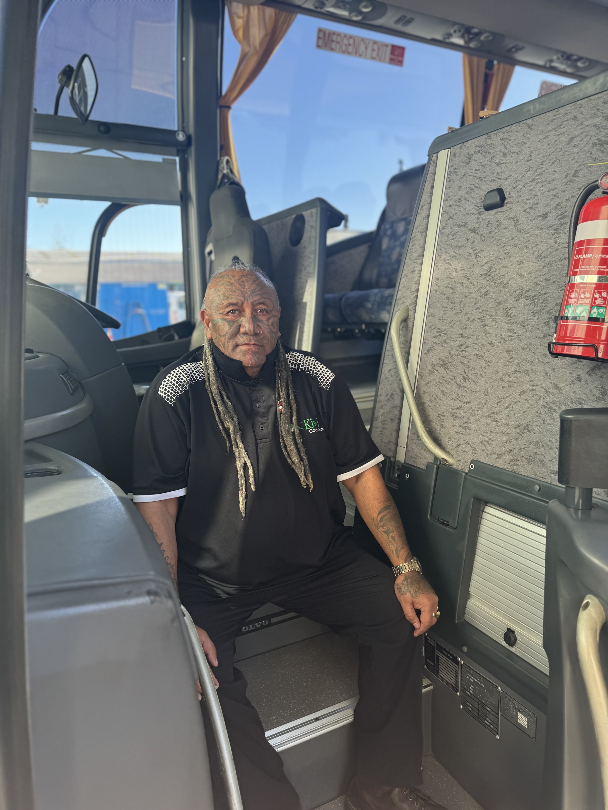 A man with tattoos and long dreadlocks sitting inside a bus near the driver's area, with a fire extinguisher mounted on the side.