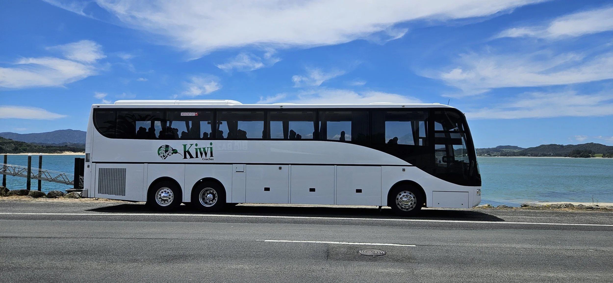 White coach bus with 'Kiwi Coaches' logo parked on the side of a road near a body of water, with mountains and blue sky in the background.
