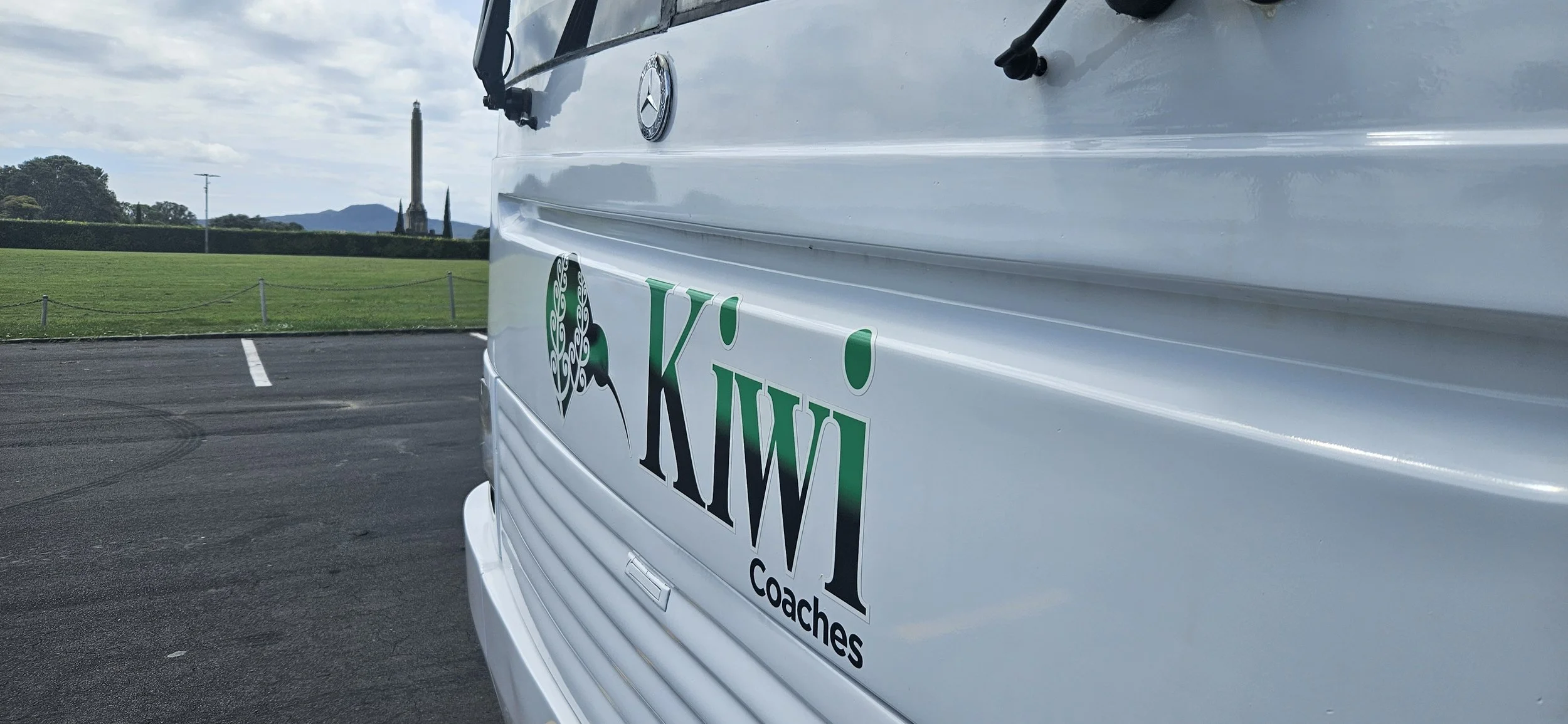 Close-up of a white vehicle with a KWI Coaches logo featuring a stylized tree with a peacock's head, parked on an asphalt lot with green grass and trees in the background.
