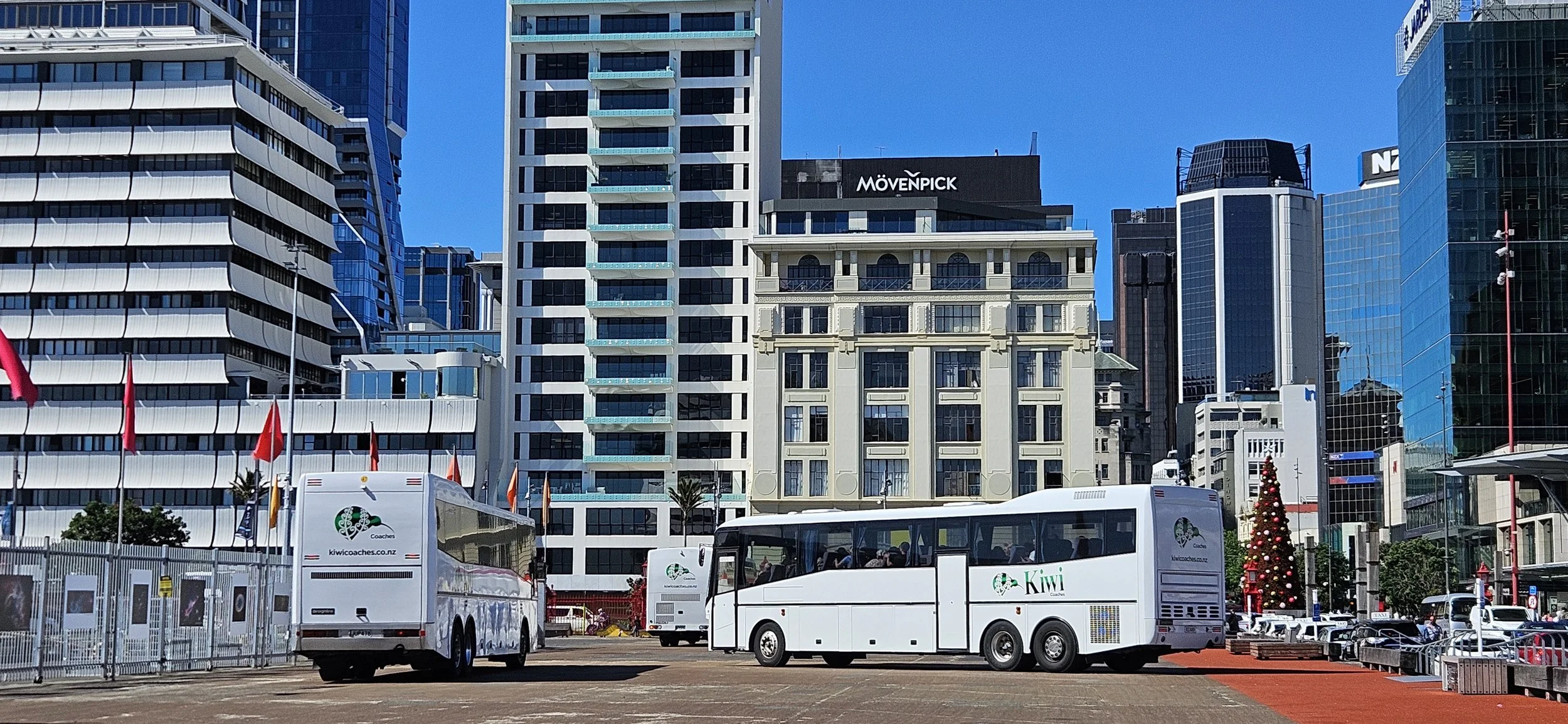 Cityscape with modern skyscrapers and a decorated Christmas tree, with white buses in the foreground in an urban area.