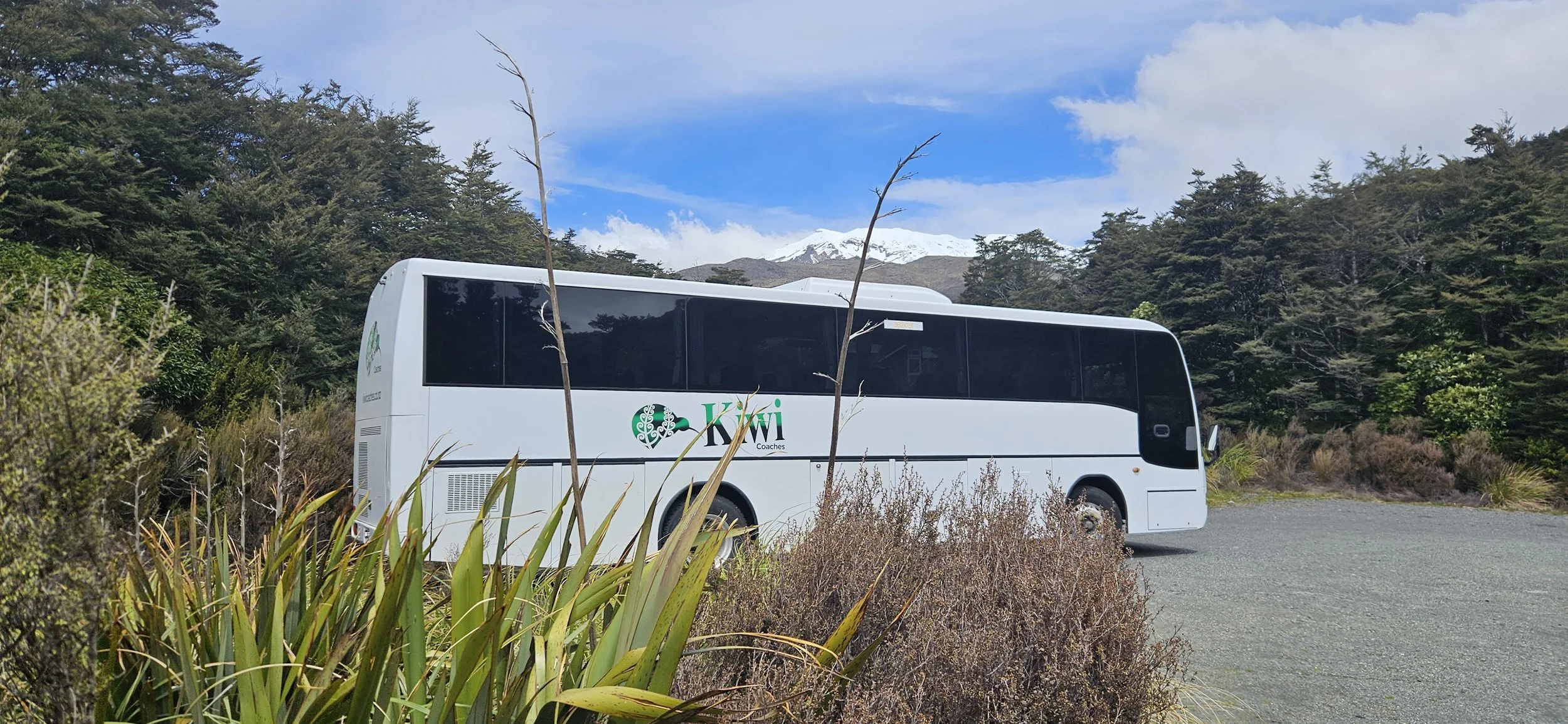 A white Kiwi Coaches bus parked on a gravel lot, surrounded by bushes and trees, with snow-capped mountains and a partly cloudy sky in the background.