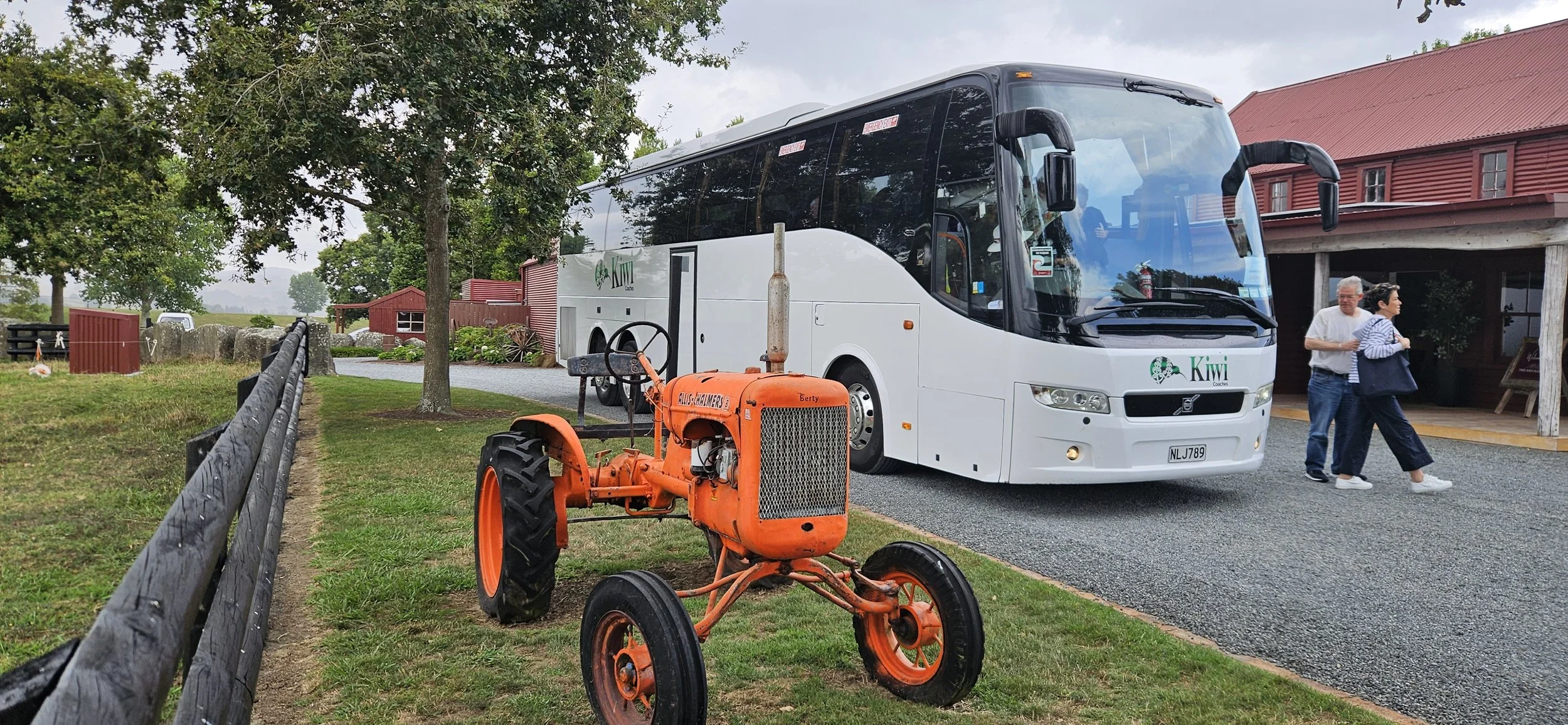 An orange vintage tractor in front of a white Kiwi coach bus parked near a building with a red roof, with two people walking nearby, on a grassy area with trees and a gravel pathway.
