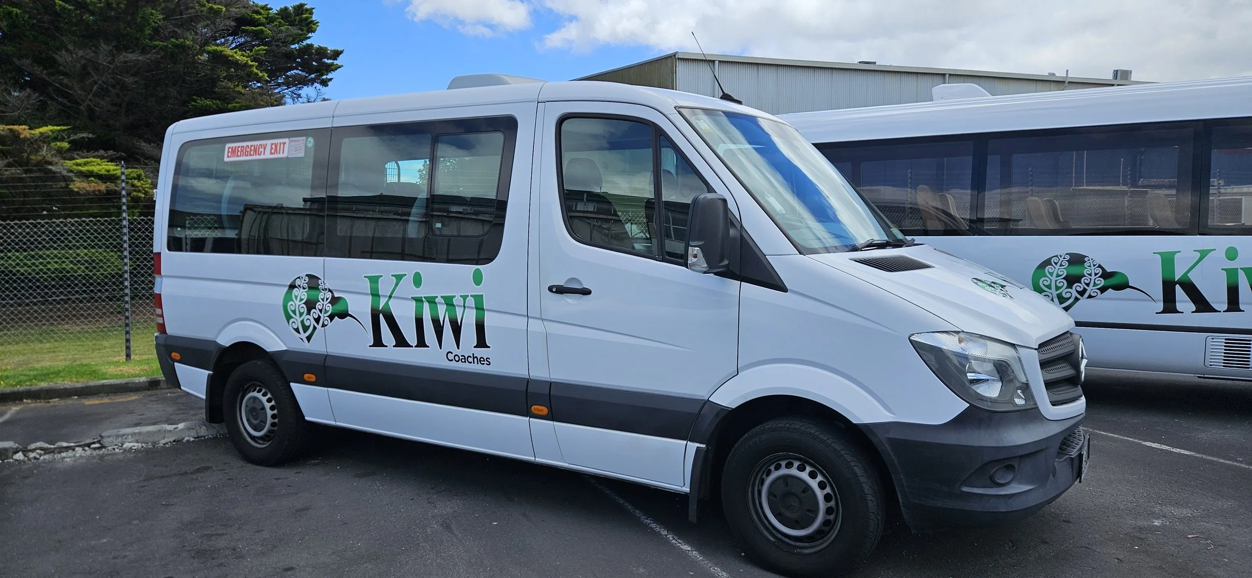 A white van with the words "Kiwi Coaches" and a kiwi bird logo on the side, parked in a parking lot with another bus visible behind it.