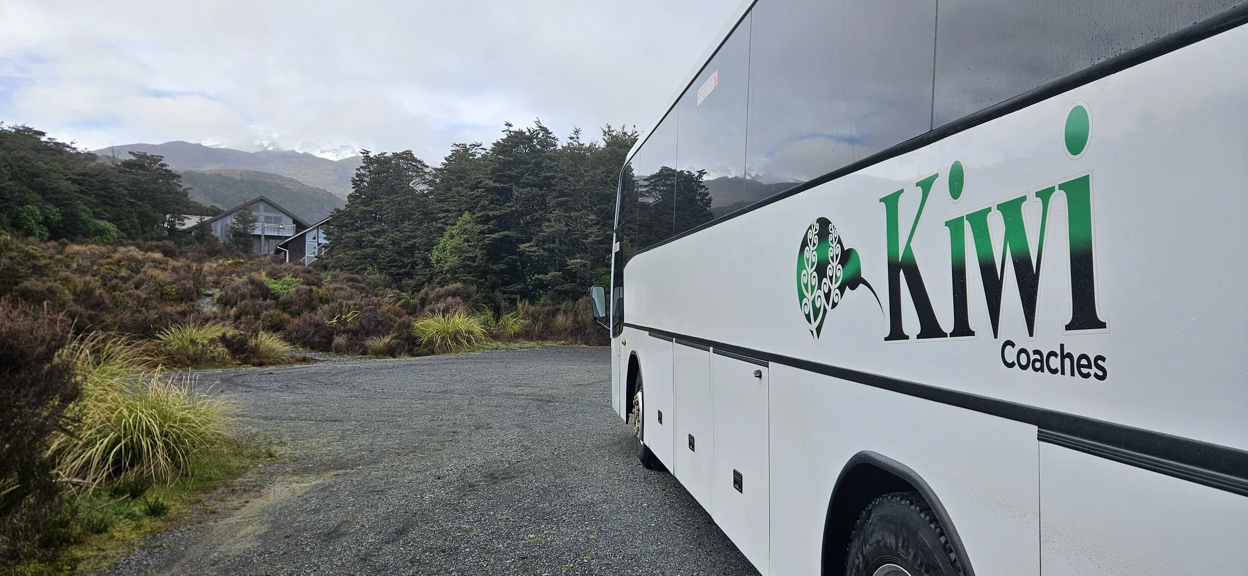 A white coach bus with the logo 'Kiwi Coaches' parked on a gravel road in a rural, mountainous area with greenery and trees, and a house or building in the background.