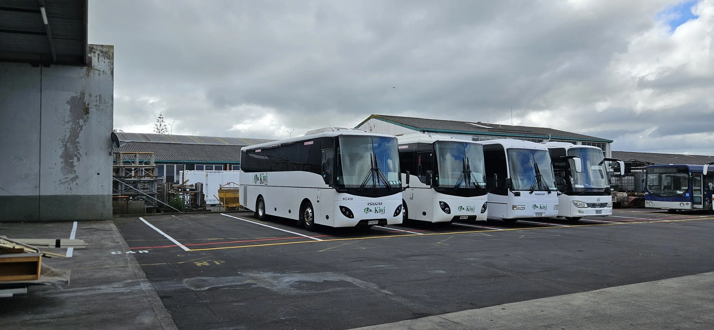 Several white buses with Kiwi markings parked in a lot on a cloudy day, with a building and cloudy sky in the background.