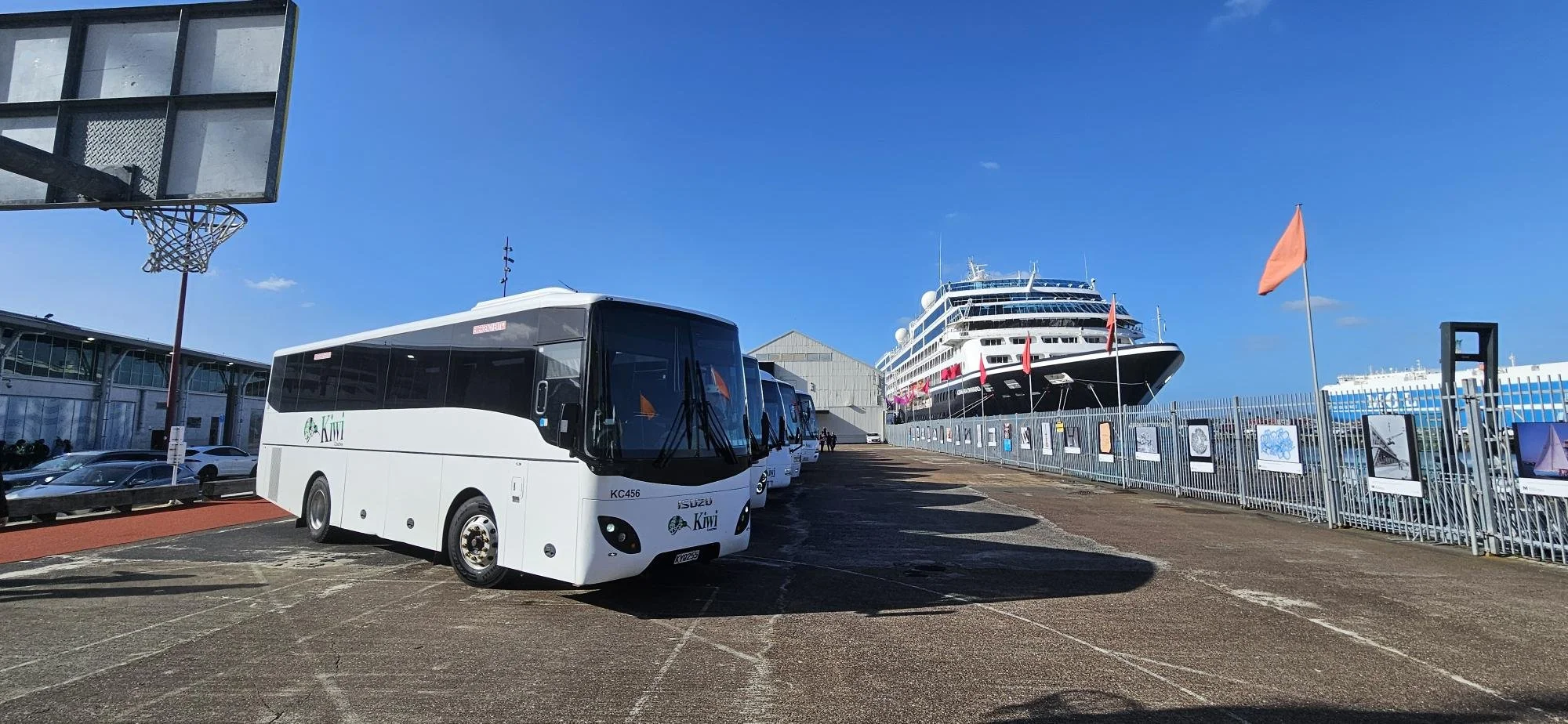 Large cruise ship docked in a port with orange flags, parked white buses with Kiwi logo, a basketball hoop in the foreground, and a fenced area displaying pictures, with a clear blue sky overhead.
