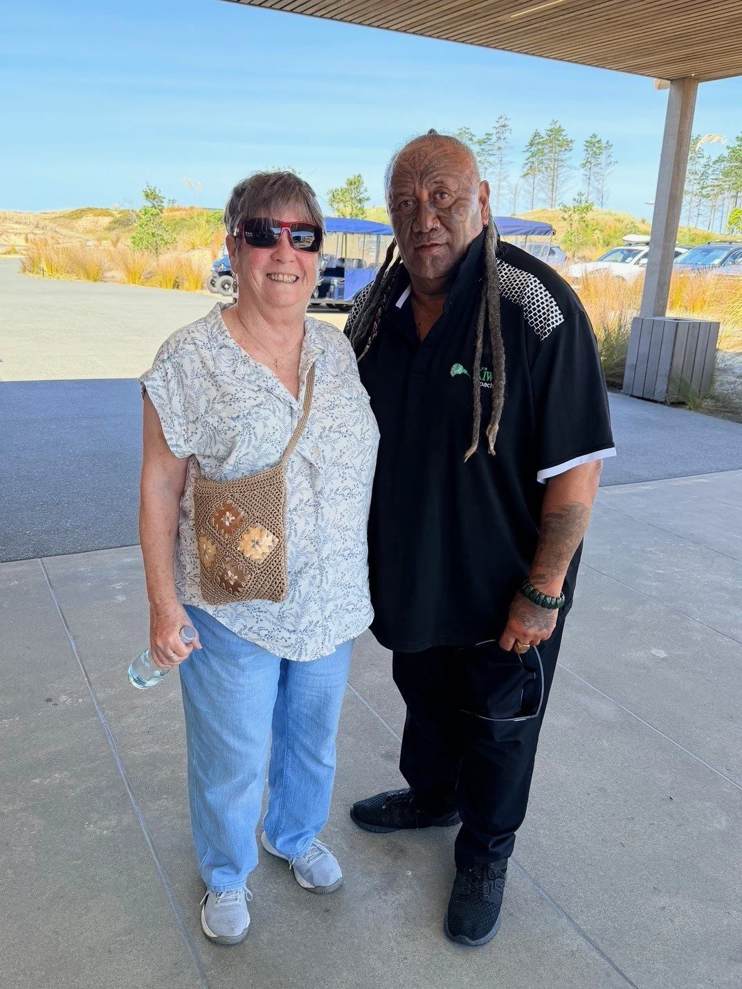 A woman wearing sunglasses, a patterned blouse, light blue jeans, and sneakers, standing next to a man with long dreadlocks, wearing a black polo shirt, black pants, and black shoes, outdoors on a sunny day.