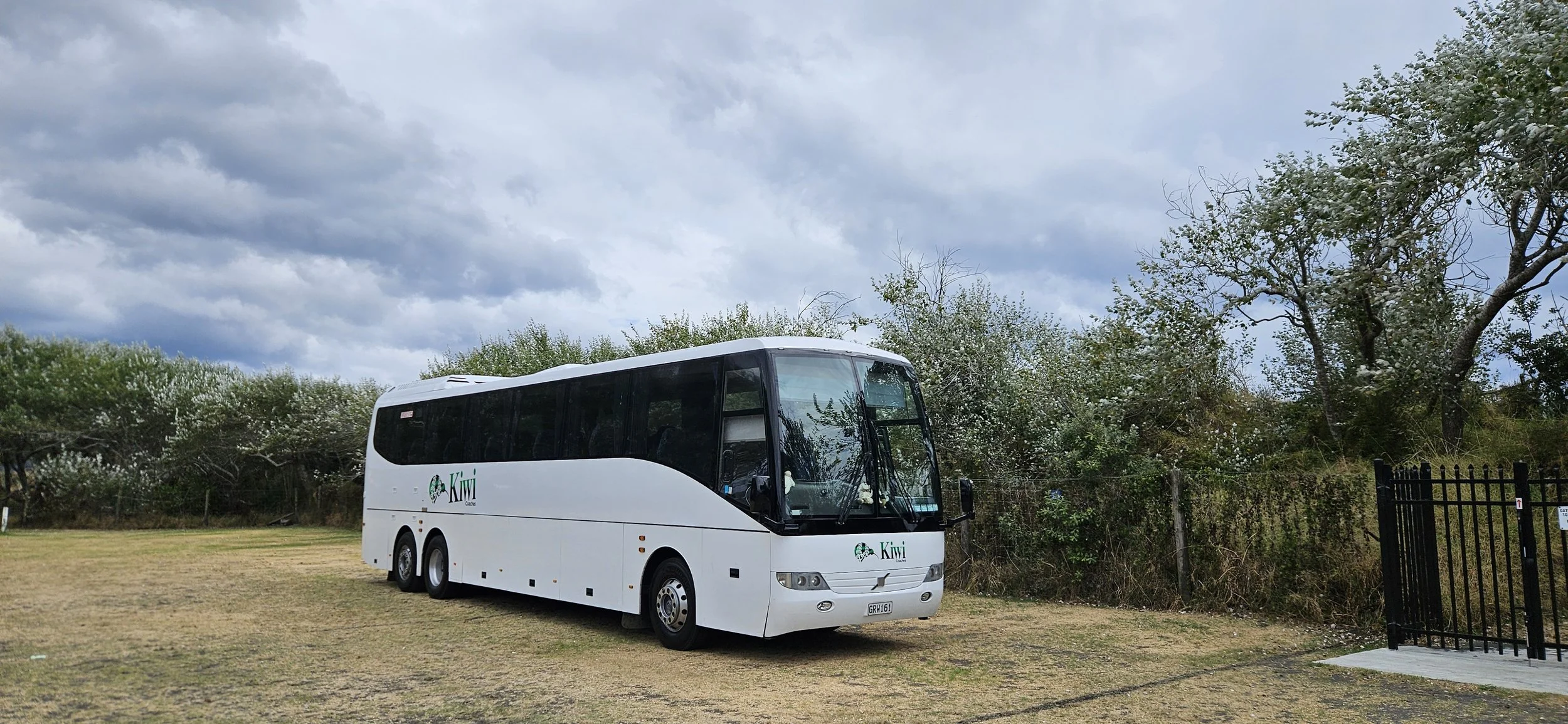 White Kiwi Tours bus parked on grassy area with trees and cloudy sky in the background.