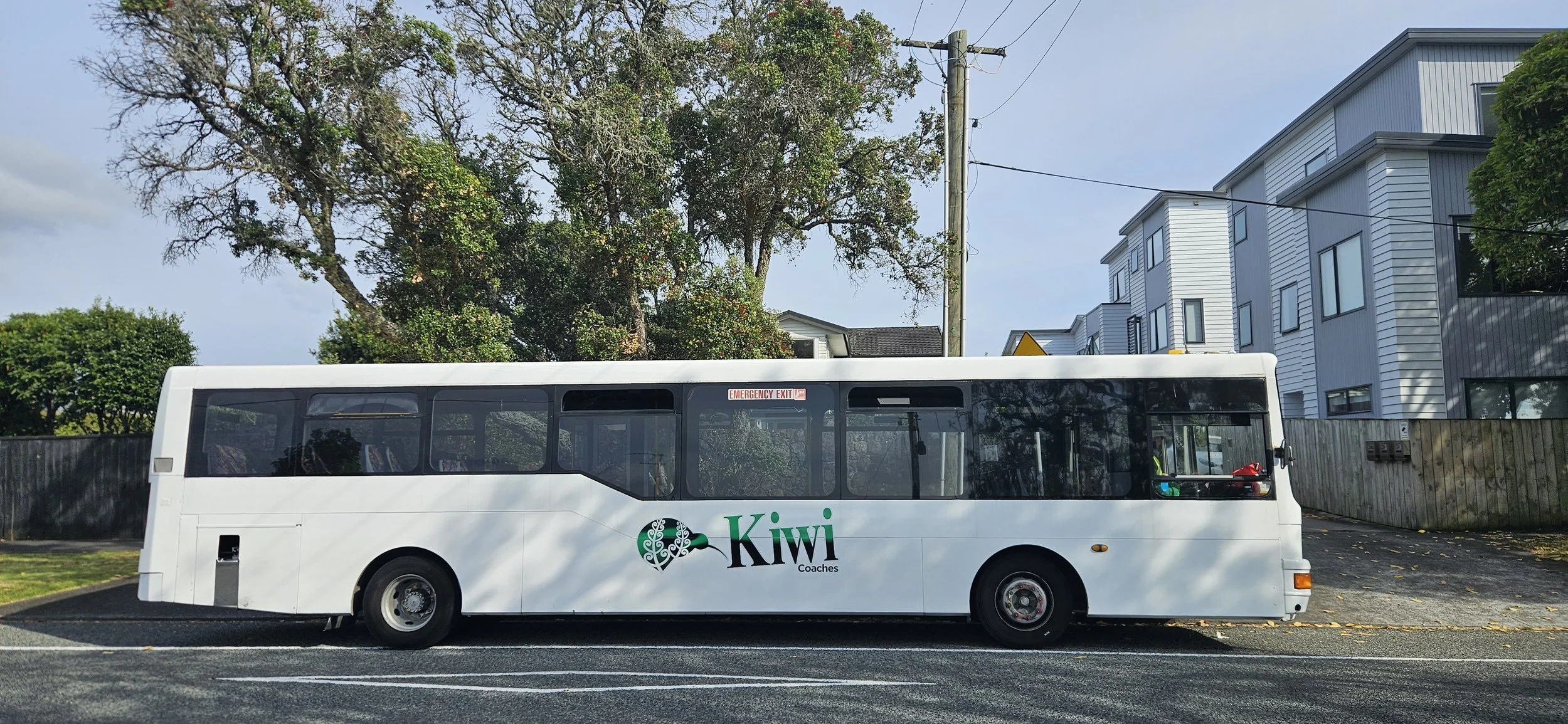 A white bus with green and black 'Kiwi Coaches' logo parked on a street with modern housing and green trees in the background.