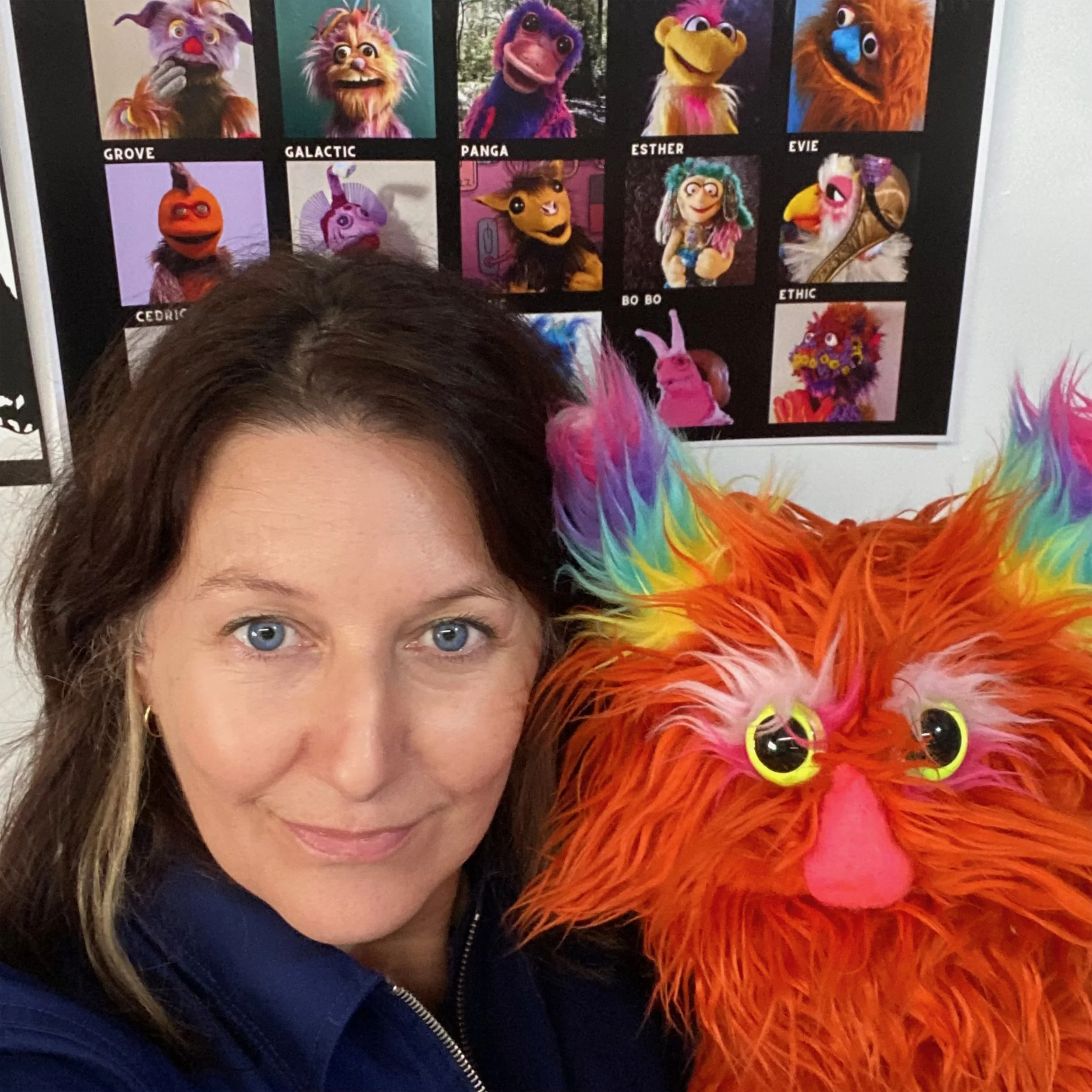 A person with dark brown hair and blue eyes posing with a colorful, furry puppet with rainbow-colored hair, bright yellow eyes, and a pink nose.