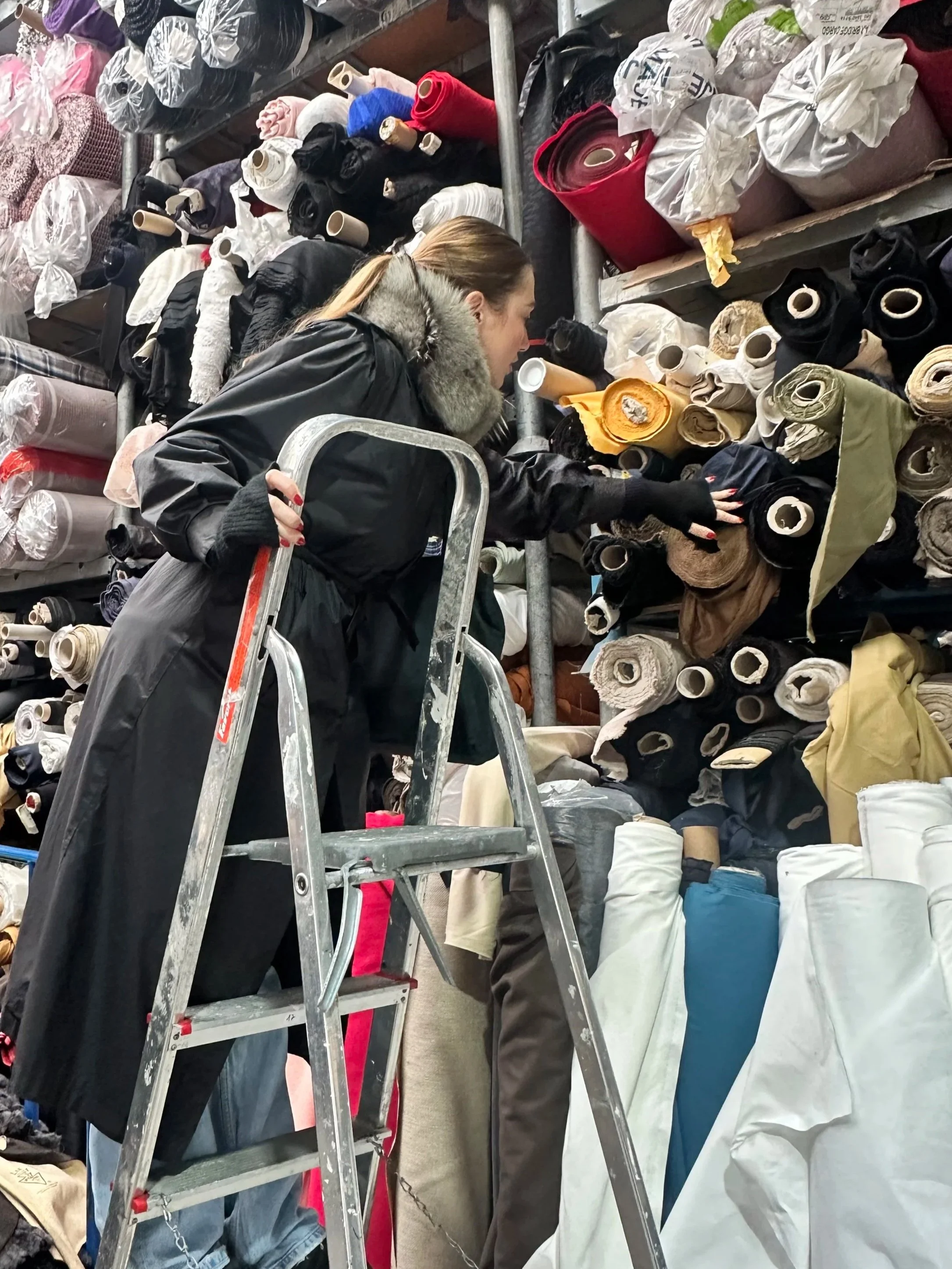Fashion designer sourcing premium deadstock fabrics from a textile warehouse filled with rolls of luxury fabrics for garment production.