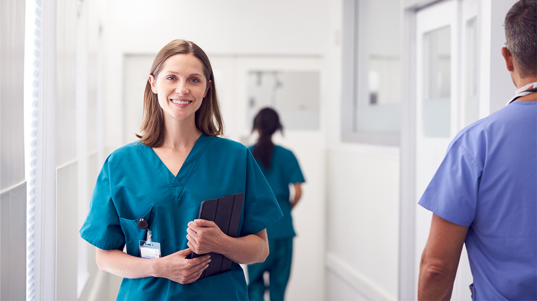 A smiling female nurse in scrubs walking through a hospital corridor.