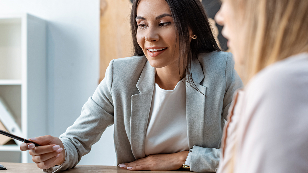 Two women having a discussion at an office table, one holding a smartphone, the other listening, professional setting.