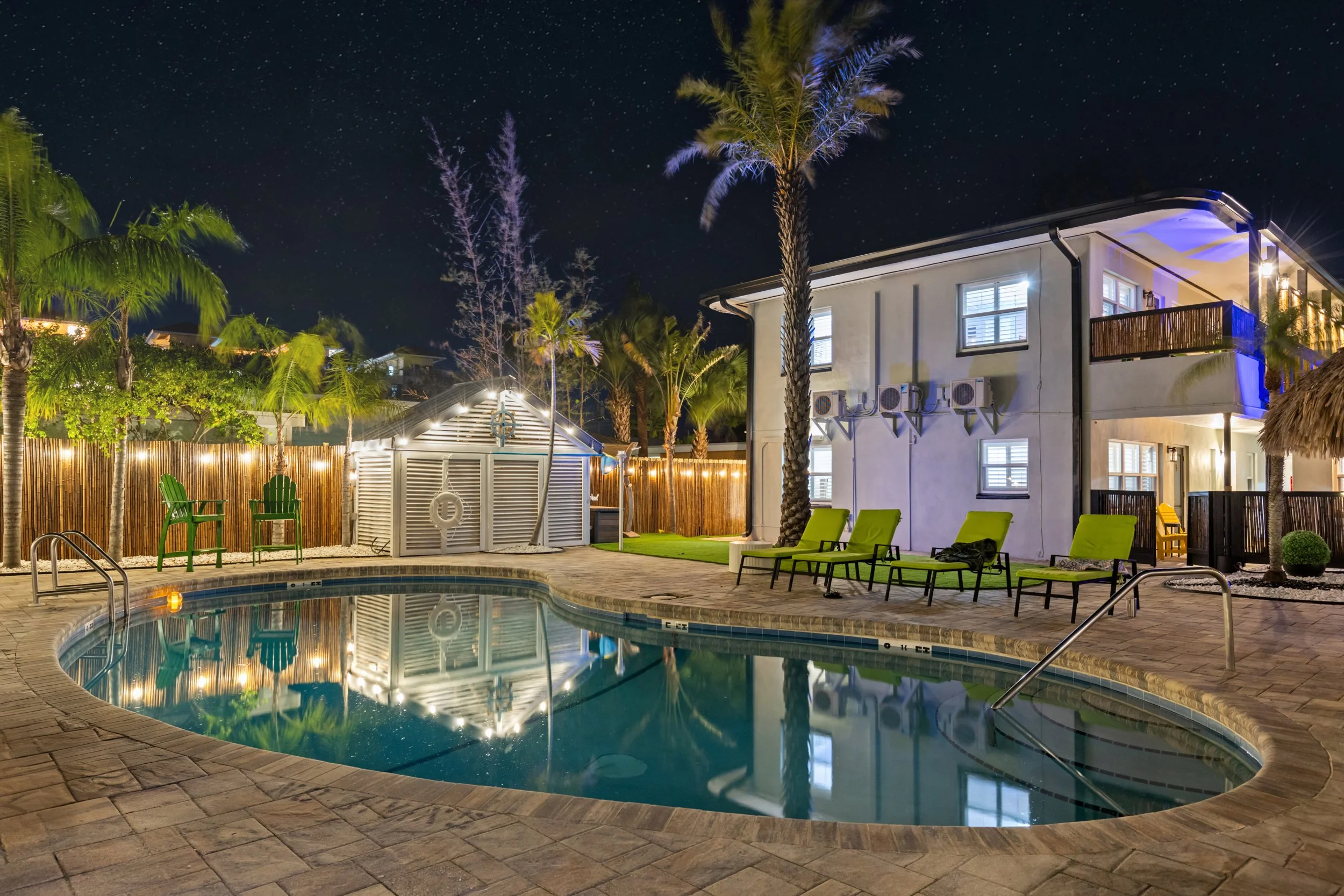Nighttime scene of a lit swimming pool surrounded by lounge chairs, palm trees, and a two-story building with illuminated windows, patio furniture, and decorative string lights.