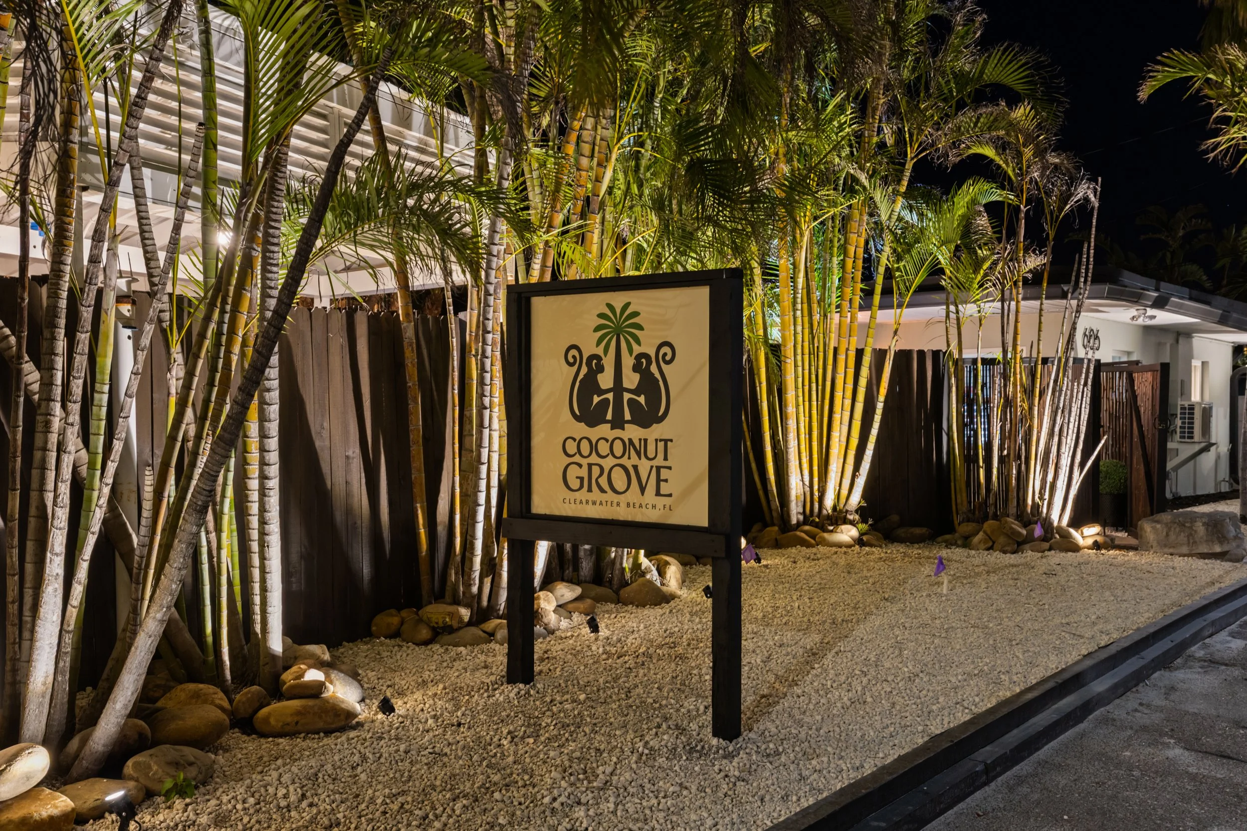 Nighttime view of Coconut Grove sign surrounded by palm trees and rocks, with a wooden fence and white building in the background, located in Clearwater Beach, Florida.