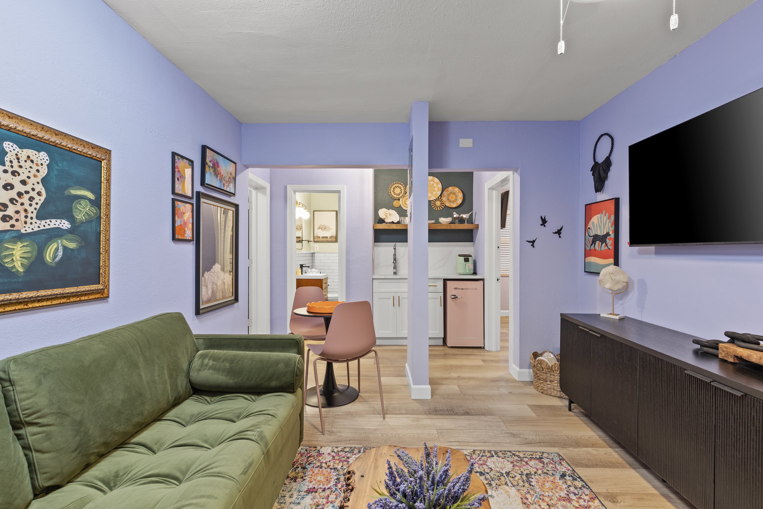 Living room with lavender walls, green sofa, pink dining chairs, artwork, a large flat-screen TV, and view into a kitchen with pastel appliances.