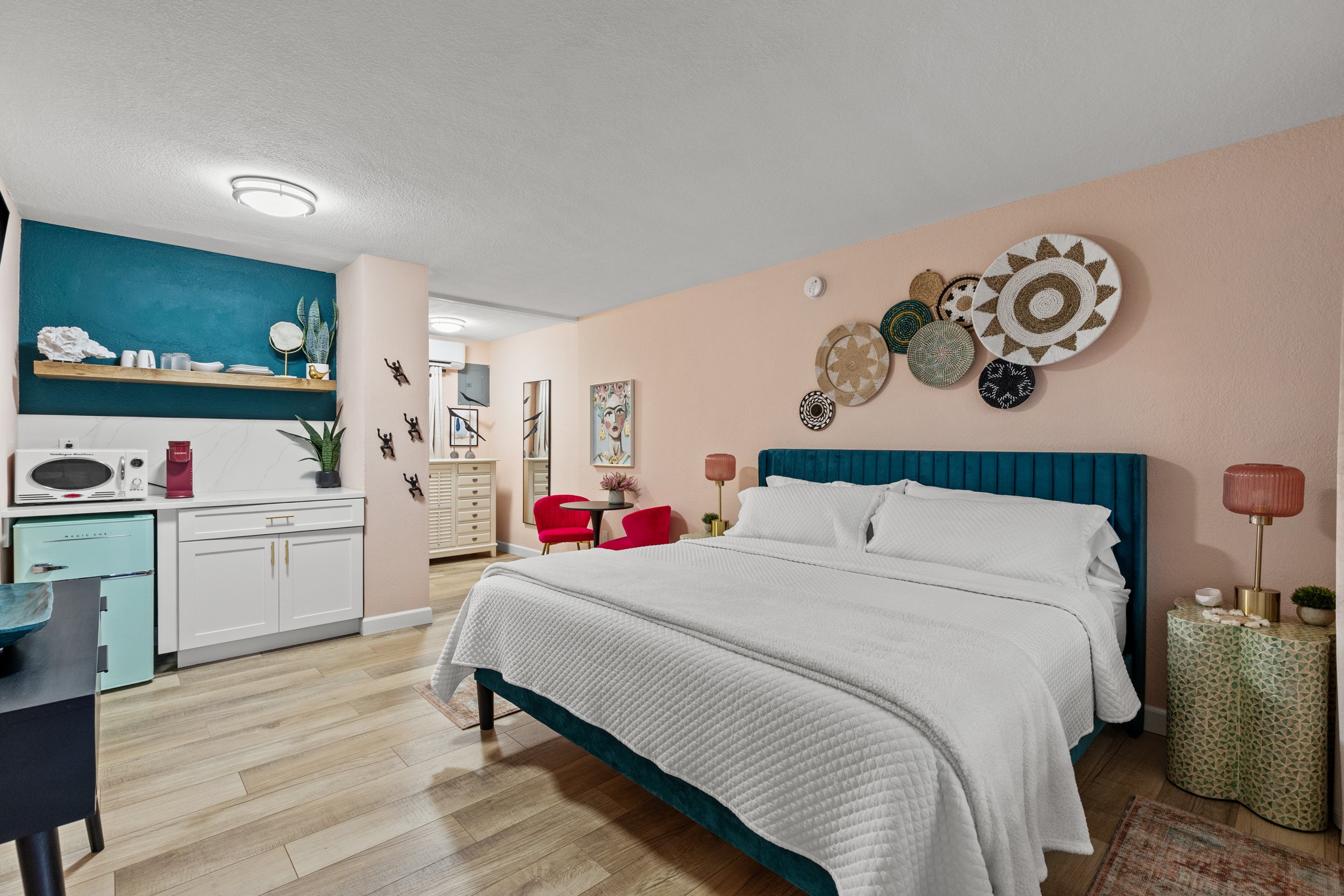 Hotel room featuring a large bed with teal headboard, pink walls with decorative baskets, a small round table with red chairs, and a kitchenette area with microwave and mini refrigerator.
